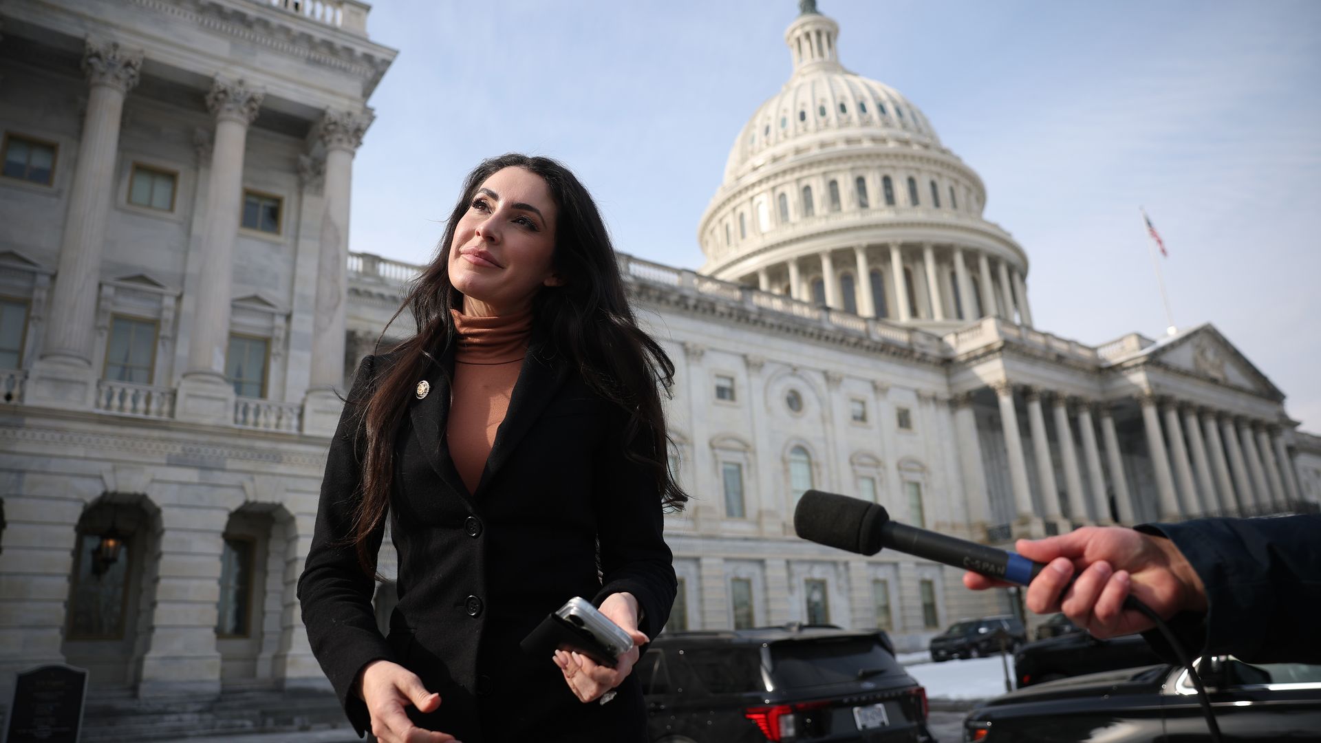 Woman in a black coat and tan turtleneck stands outside the U.S. Capitol as a reporter holds a microphone toward her; cars are parked and the clear sky forms the backdrop.