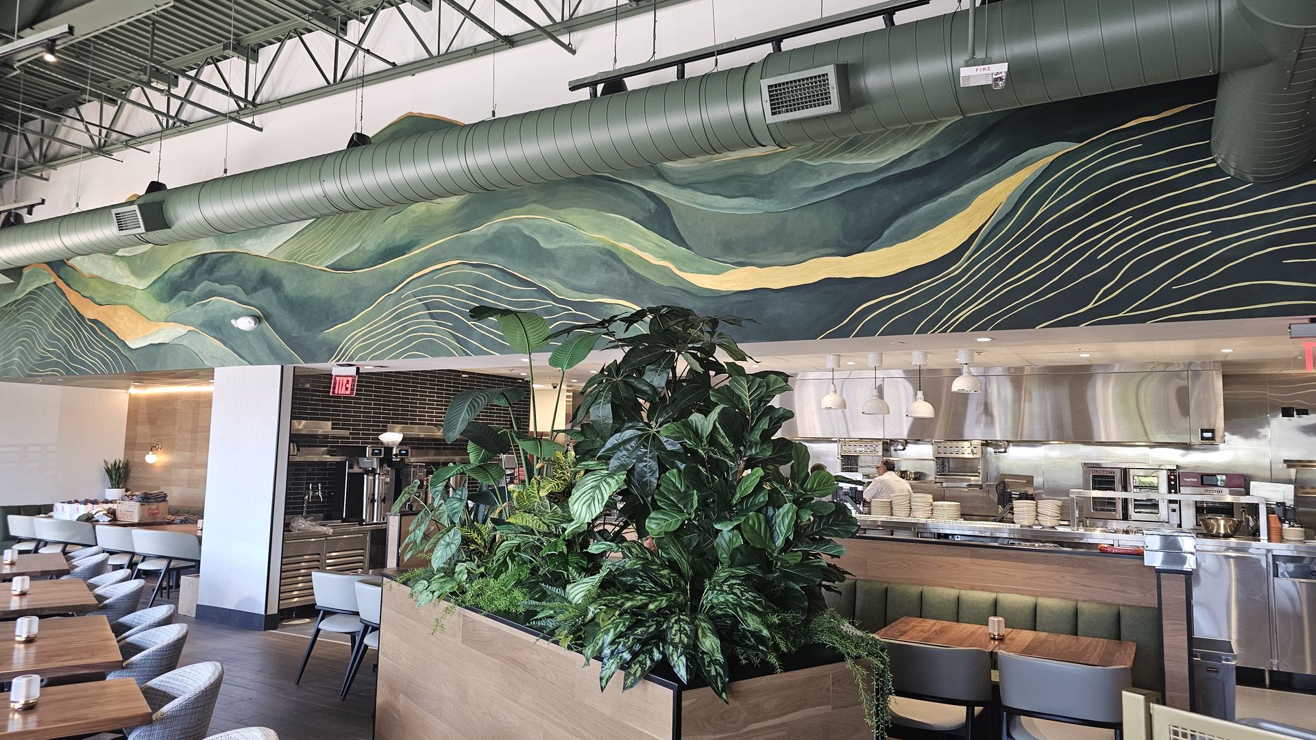 Modern cafe interior with a mural of green hills and gold waves, exposed ductwork above, lush plants, and wooden tables and chairs near an open kitchen.