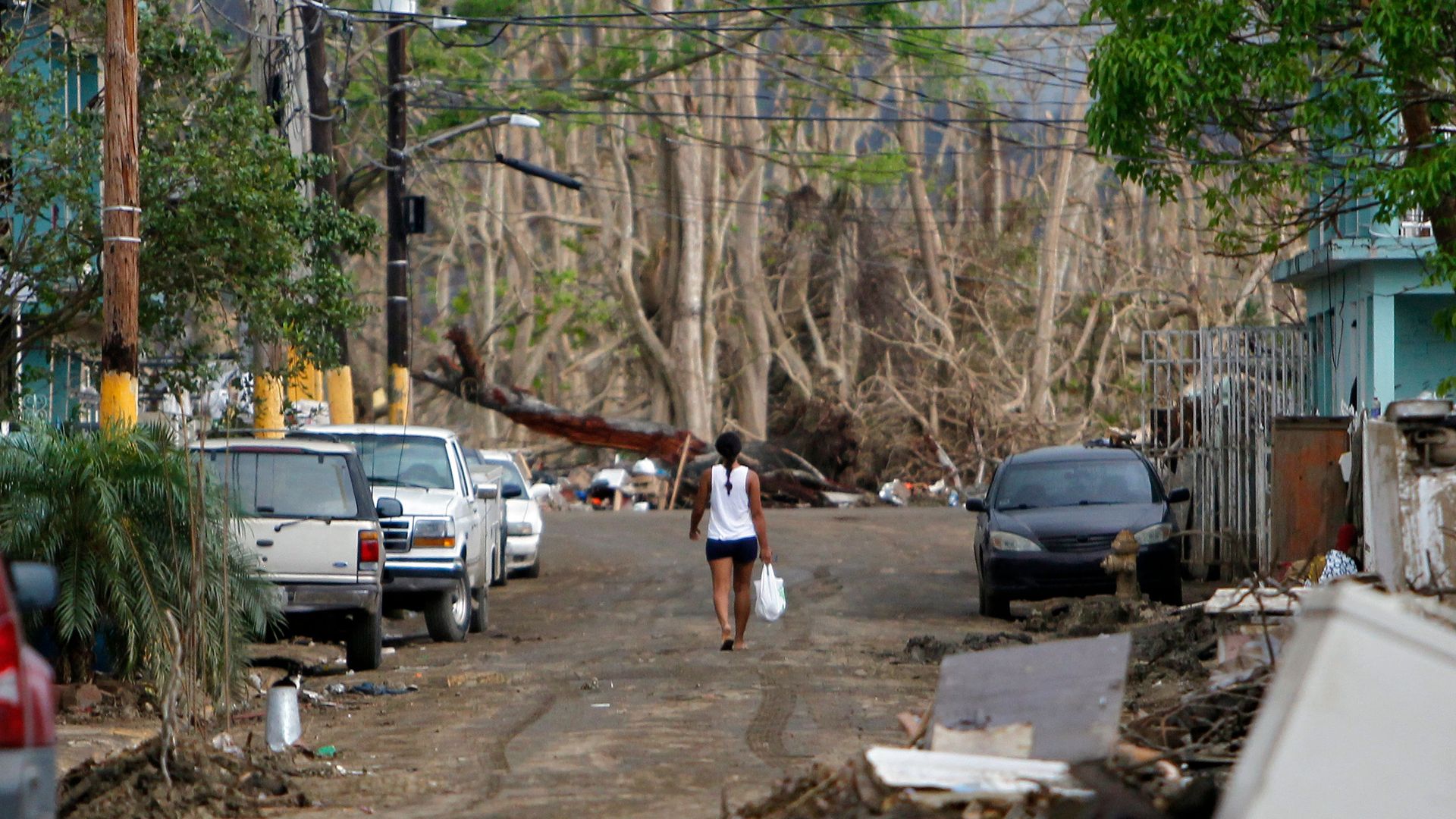 A woman in a white tank top walks down the middle of a dirt road with cars on either side.