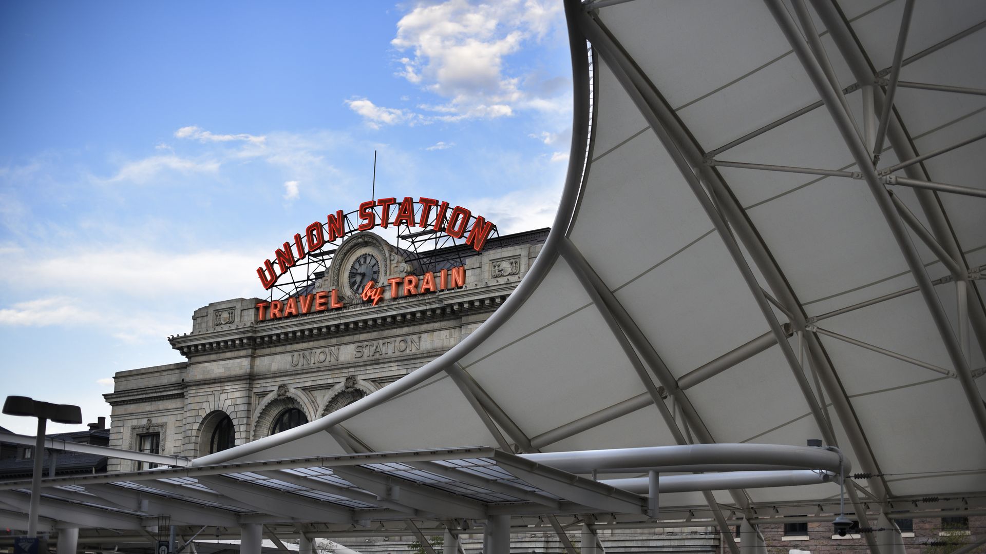 A gray, historic building with the words UNION STATION TRAVEL BY TRAIN in bright orange letters is seen in the background, while a massive metal roof is visible in the foreground to the building's right. 