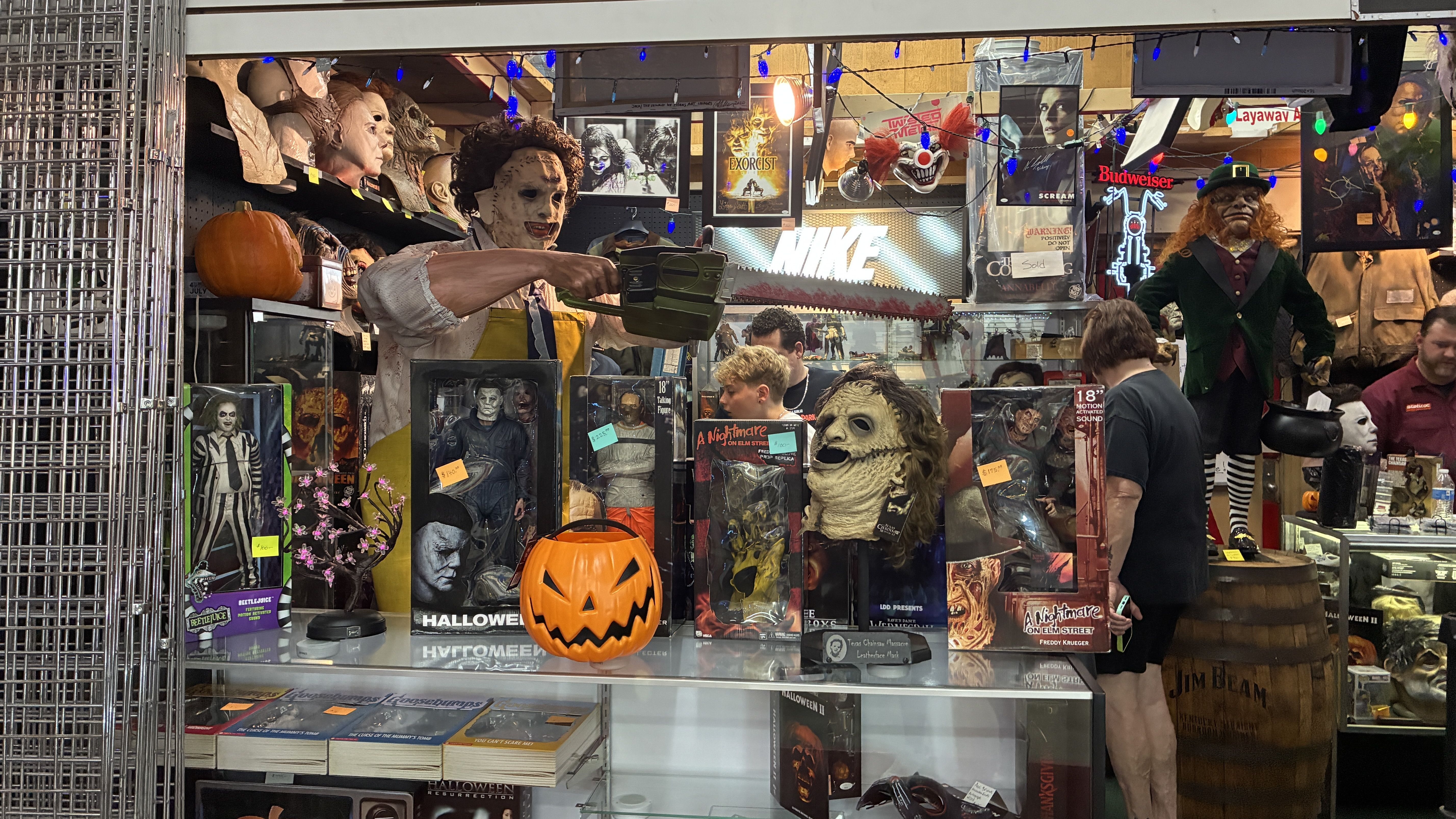 Halloween prop shop window with horror figures, a life-size masked man wielding a chainsaw, a carved pumpkin, and posters for "Exorcist" and "A Nightmare on Elm Street," plus string lights.