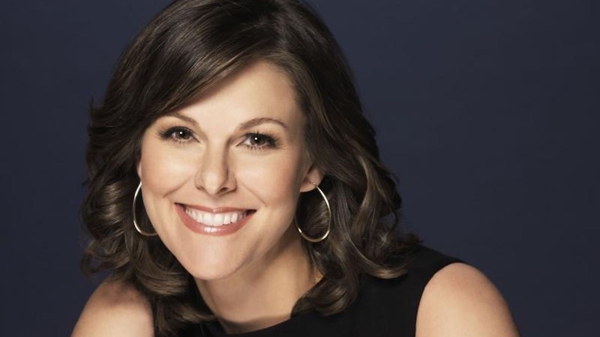 Smiling woman with shoulder-length brown hair and hoop earrings wearing a sleeveless black top against a dark background