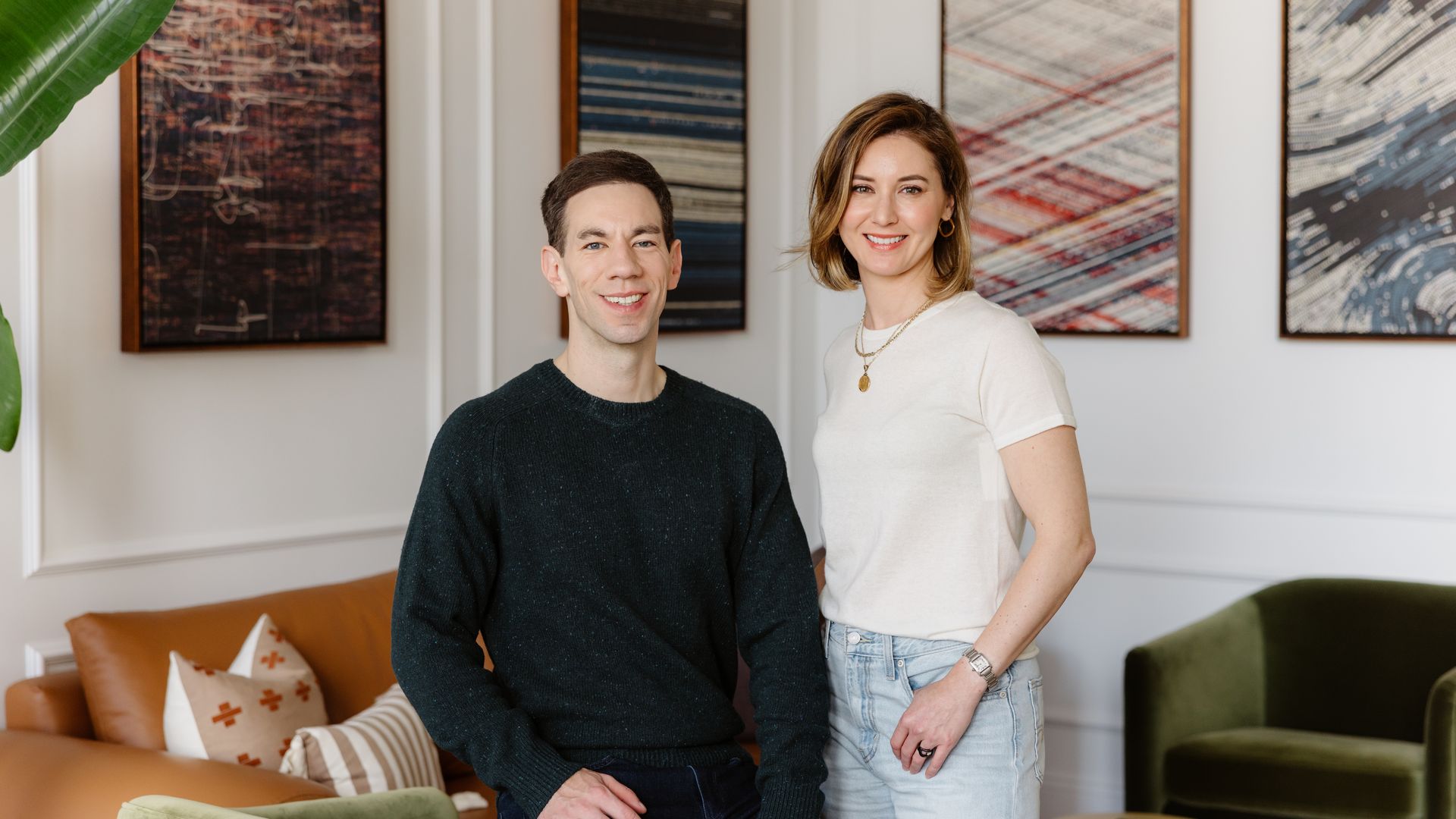 Two people smile in a bright living room with abstract art on the wall. A man in a dark sweater sits near a green chair, while a woman in a white top and jeans stands beside him.