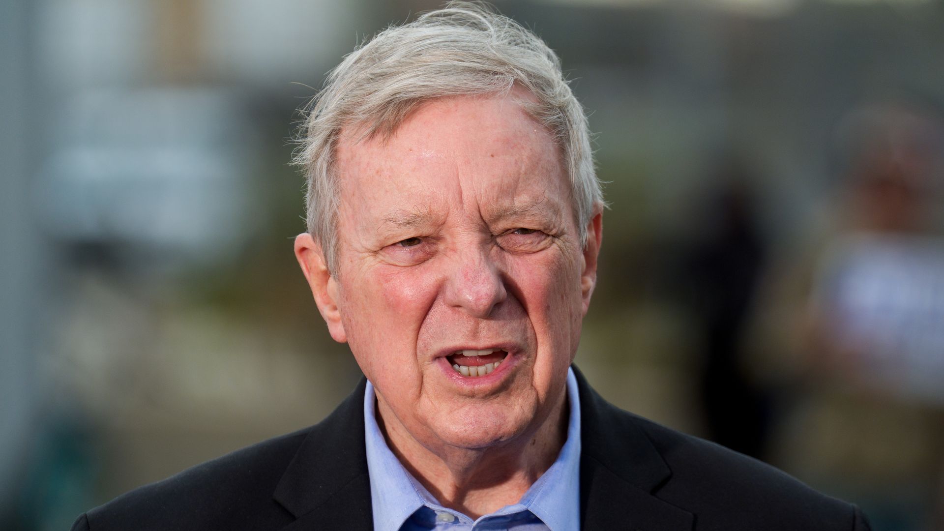 Older man with gray hair wearing a black blazer and light blue shirt, speaking outdoors with a blurred background of people and buildings.