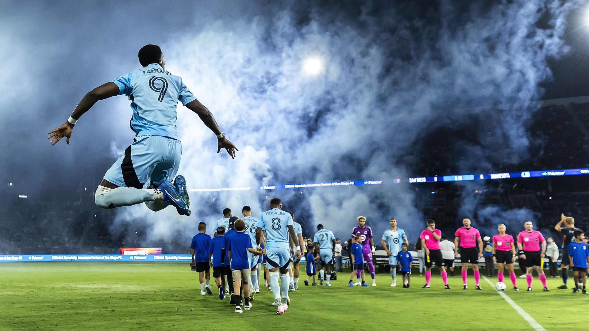 Soccer player in light blue jersey with number 9 jumps in the air, celebrating as teammates and referees in pink shirts line up on a green field under smoky night sky lights.