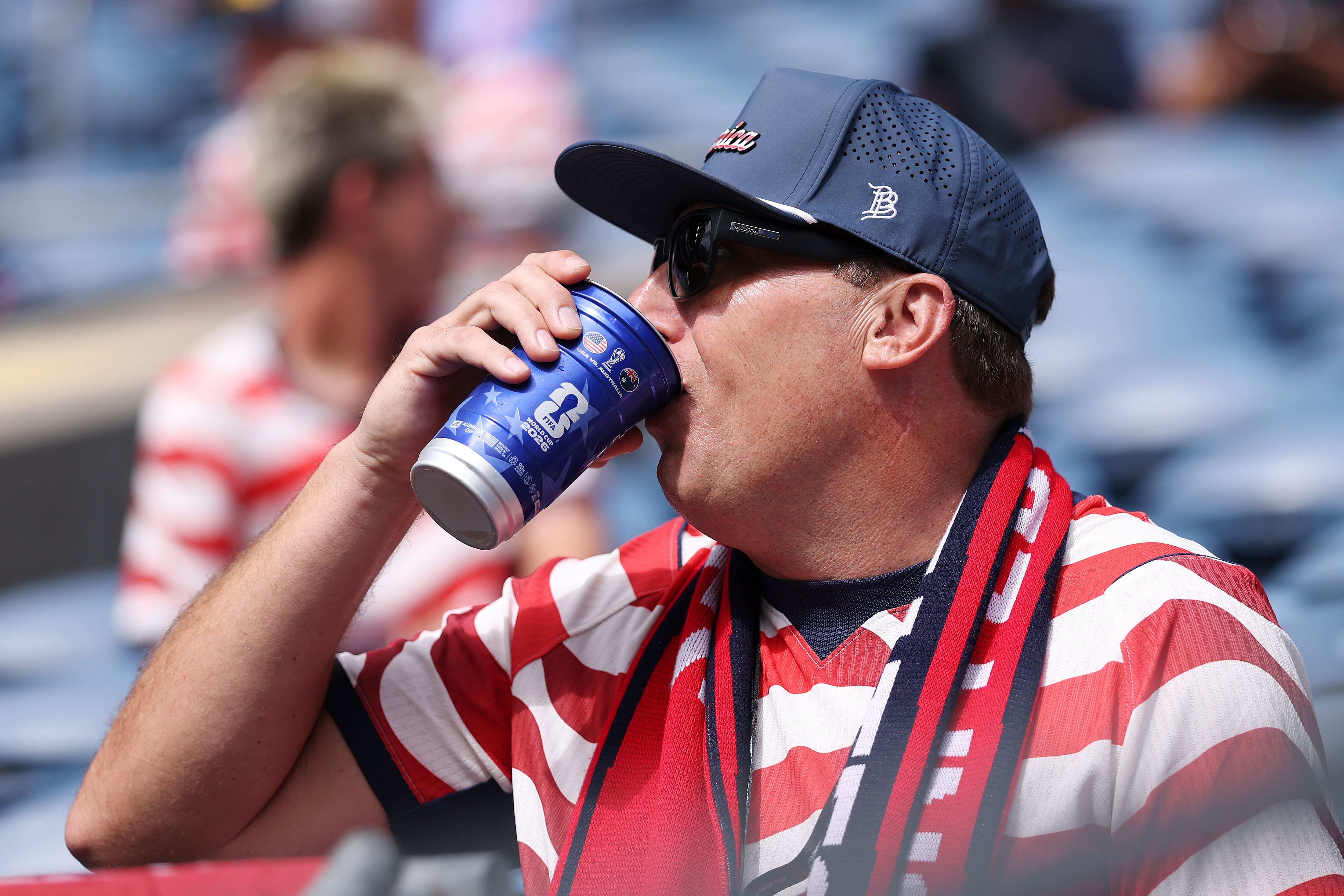 A man wearing a blue ballcap, sunglasses and USA jersey with red and white stripes drinks from a blue cup while looking off camera. In the background, other fans in can be seen in similar jerseys.