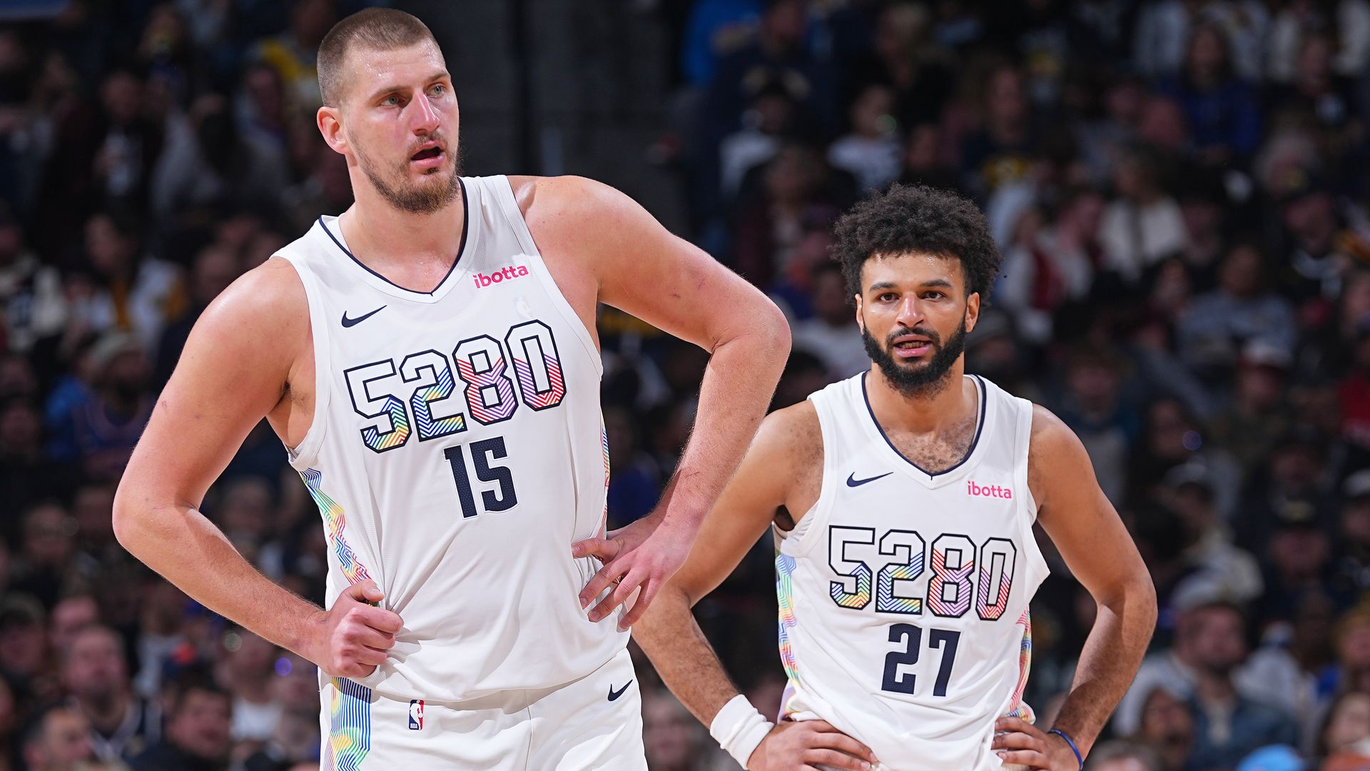 Two men in white basketball jerseys with the numbers 5280 on them stand while holding their hips and looking onward. 