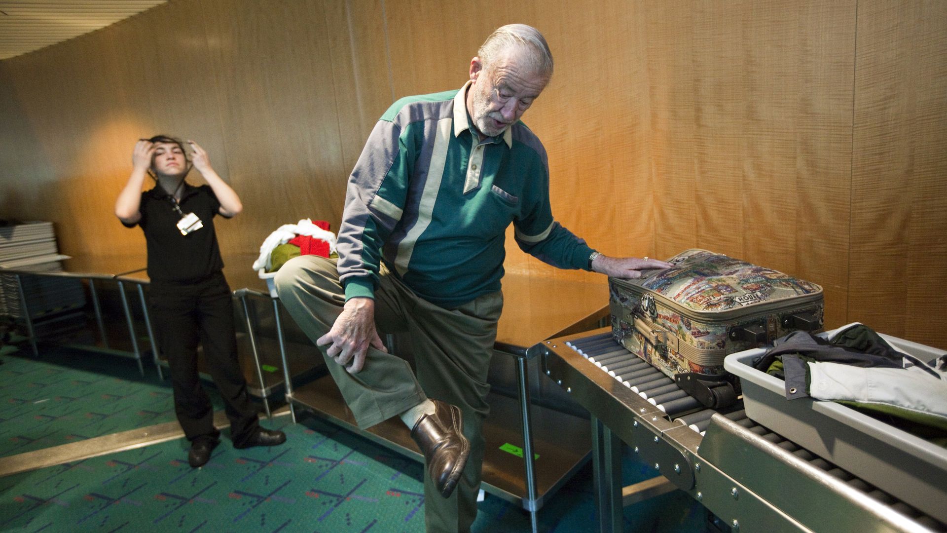 An older man in a green and gray sweater lifts his leg, placing a hand on his shoe at a security checkpoint with a conveyor belt holding a patterned suitcase and a bin of clothes. A security officer stands nearby.