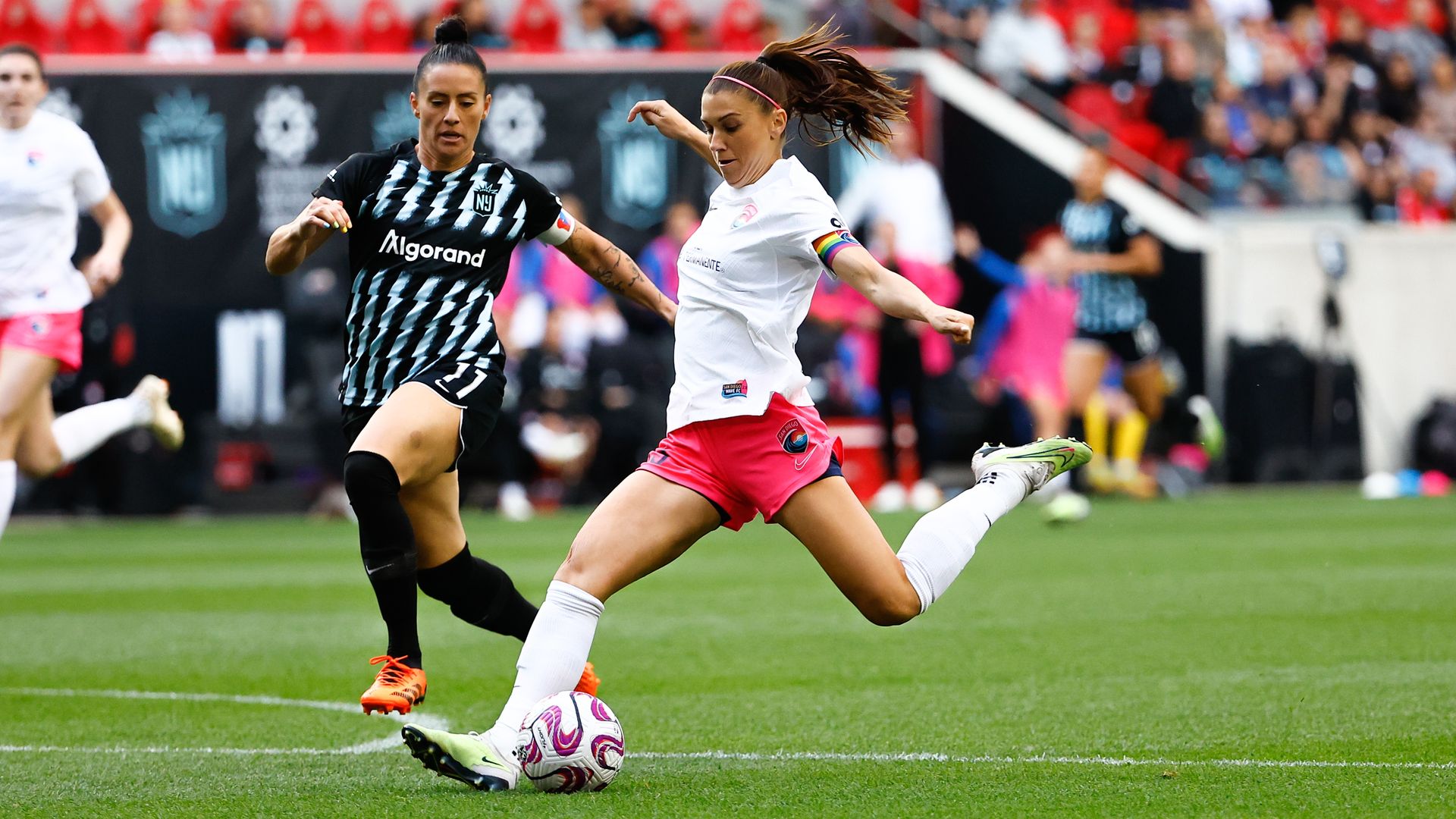 Photo of Alex Morgan about to kick a soccer ball during a match as an opponent runs alongside her