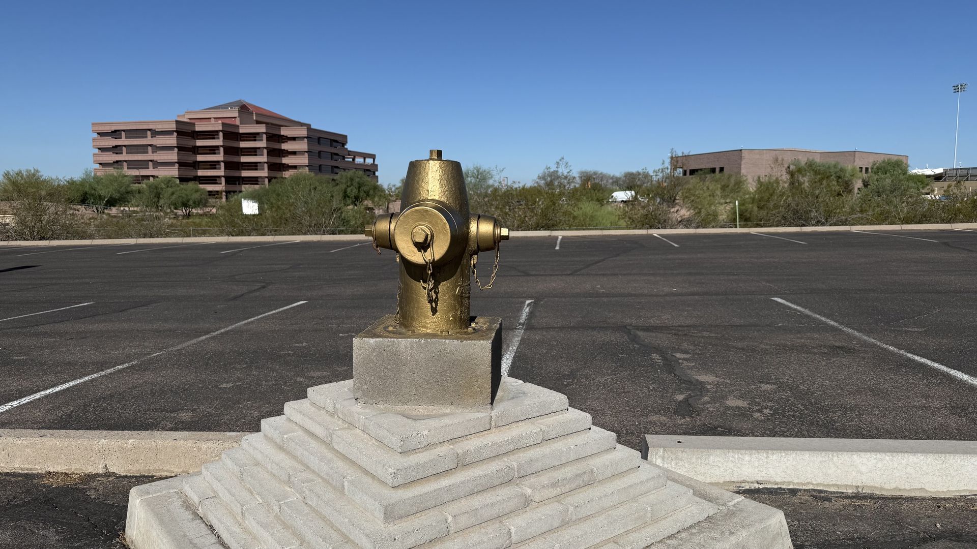 Gold-painted fire hydrant on tiered concrete base in an empty parking lot with clear blue sky and office buildings in the background.
