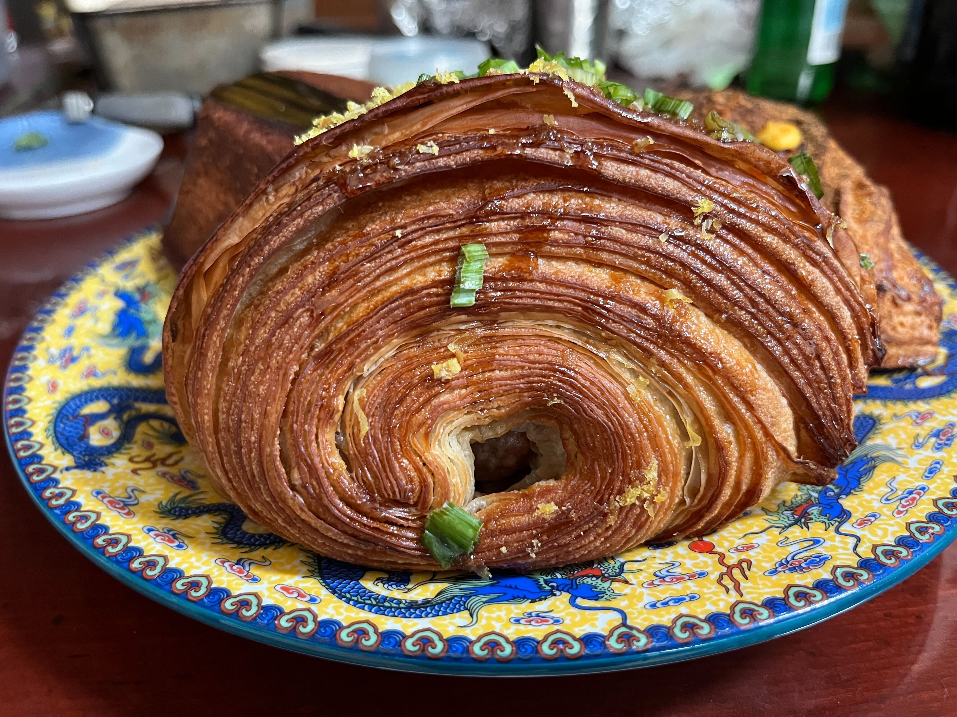 Close-up of a spiral-shaped golden brown pastry with crispy layers, garnished with green herbs, on a vibrant yellow plate decorated with blue dragons and red accents.
