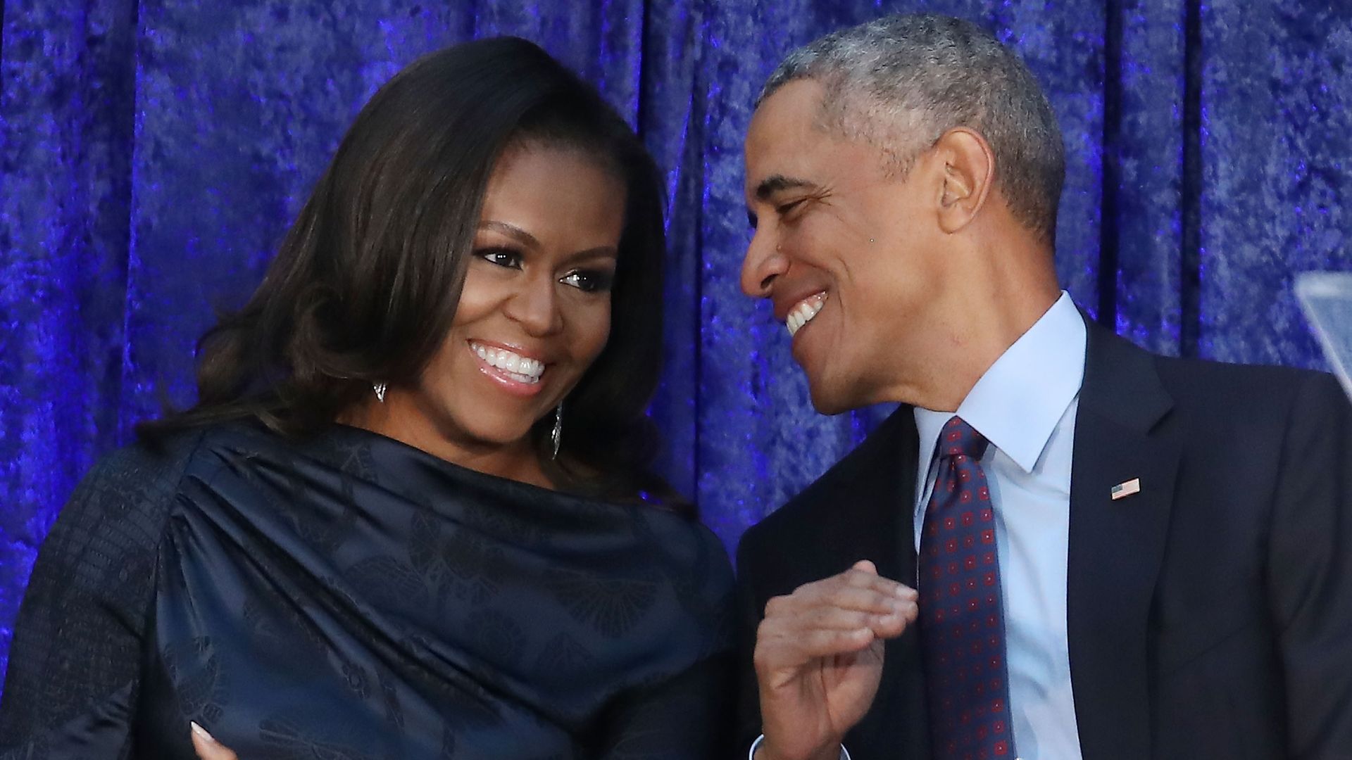 Barack and Michelle Obama sitting down next to each other and leaning toward each other laughing. 