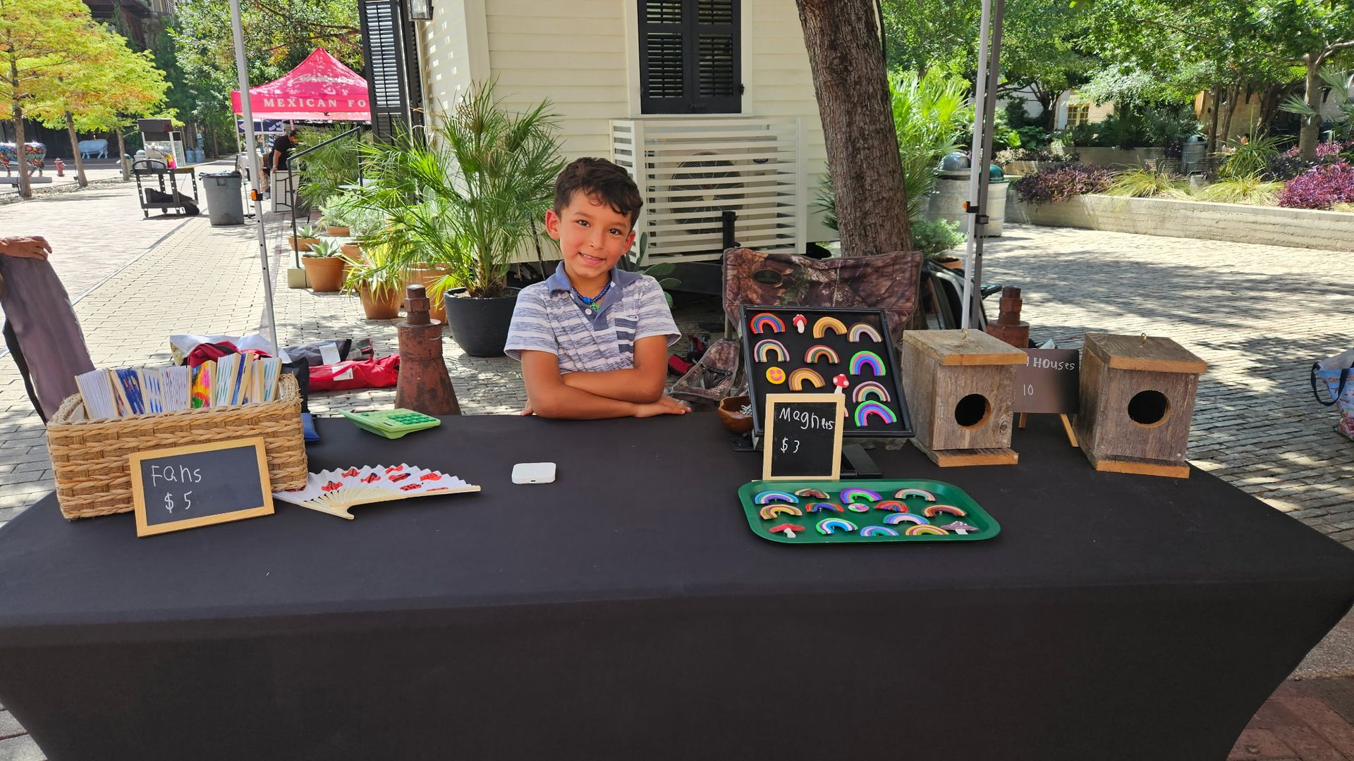 Young boy sells handmade colorful fans, rainbow magnets, and wooden birdhouses at outdoor market table with black cloth on sunny day.