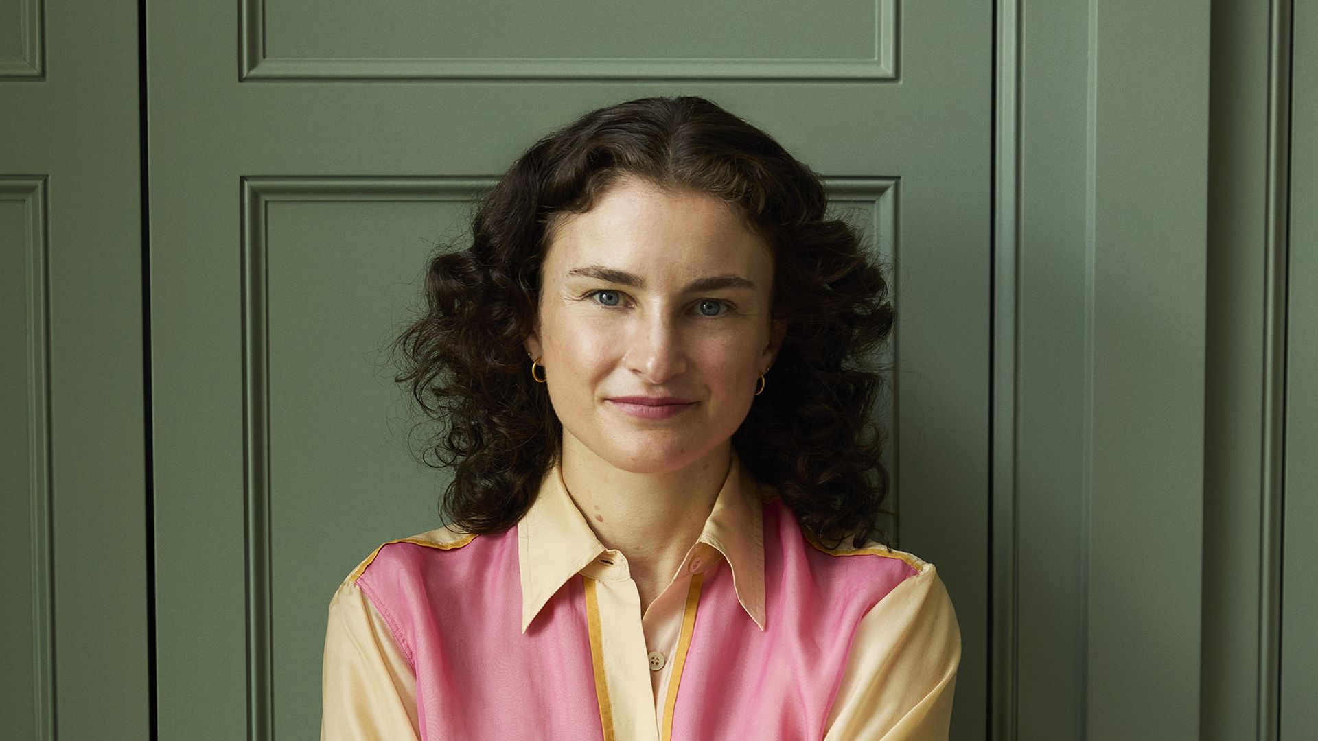 Woman with curly dark hair wearing a beige shirt with a pink vest and blue jeans, standing with arms crossed against green paneled doors with gold handles.
