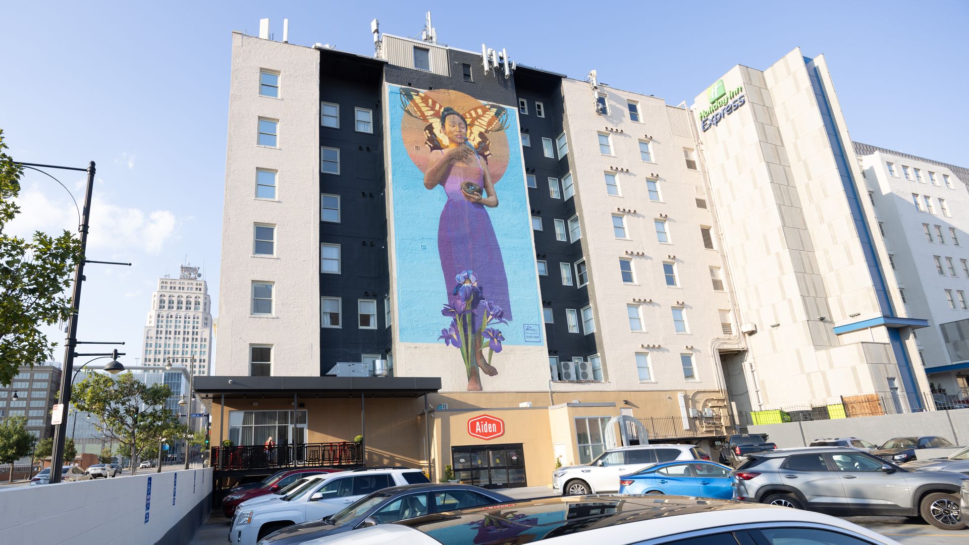 City street with a tall building mural: a woman in a purple dress with butterfly wings and an orange halo; iris flowers at the bottom. Holiday Inn Express on the right, with many parked cars.