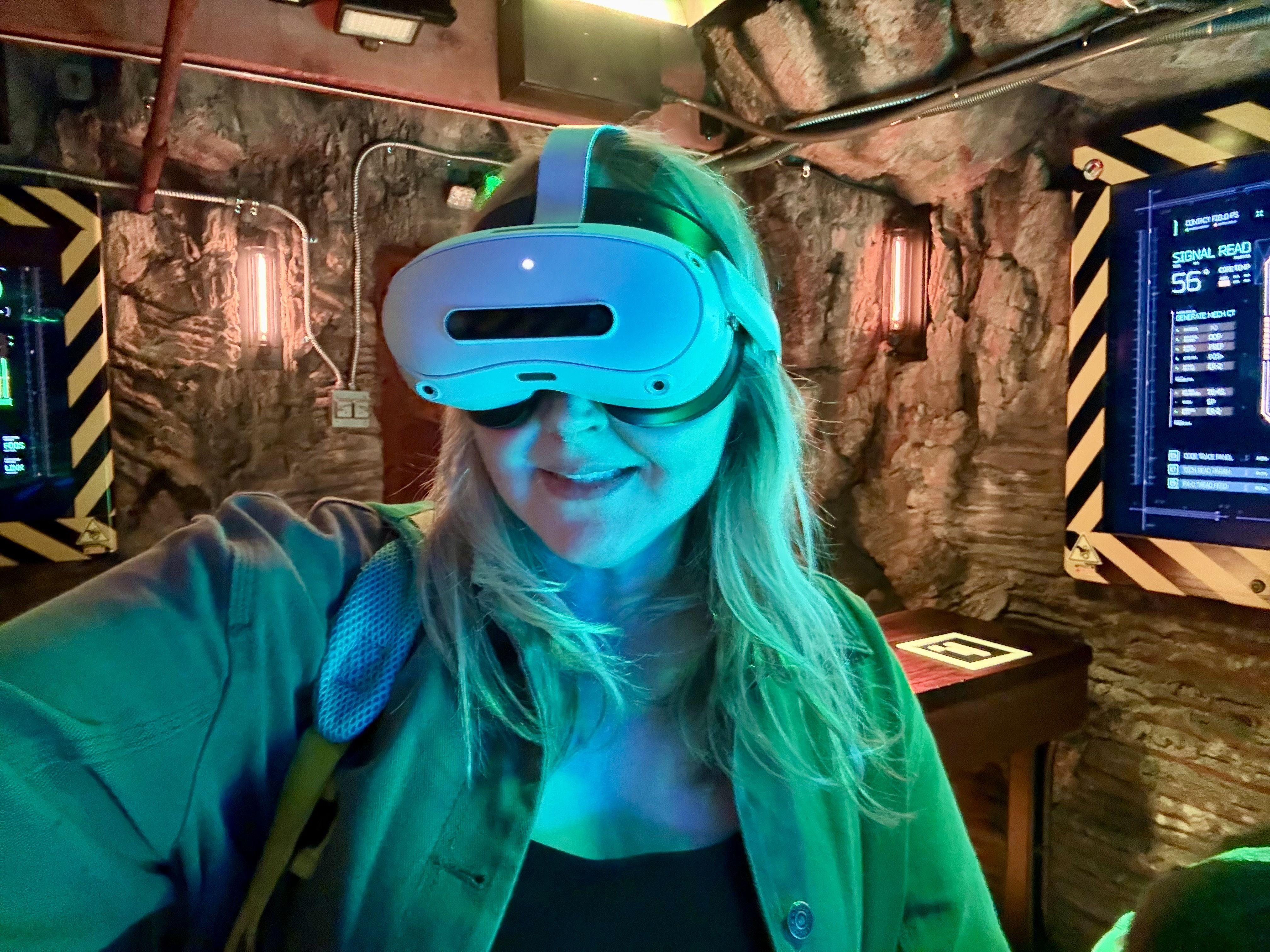 Selfie of a person wearing a large white and blue VR headset in a cave-like setting with yellow-and-black hazard stripes, exposed wiring, and a digital display reading "Signal Read" in the background.