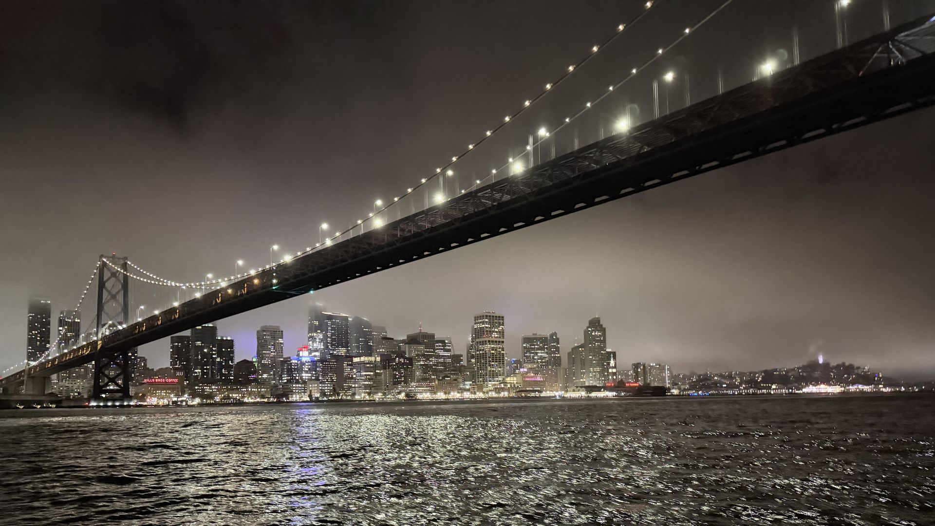 Night image of a lit suspension bridge crossing a foggy bay, with city skyscrapers glowing in the background. Rows of bright white lights line the cables and tower.