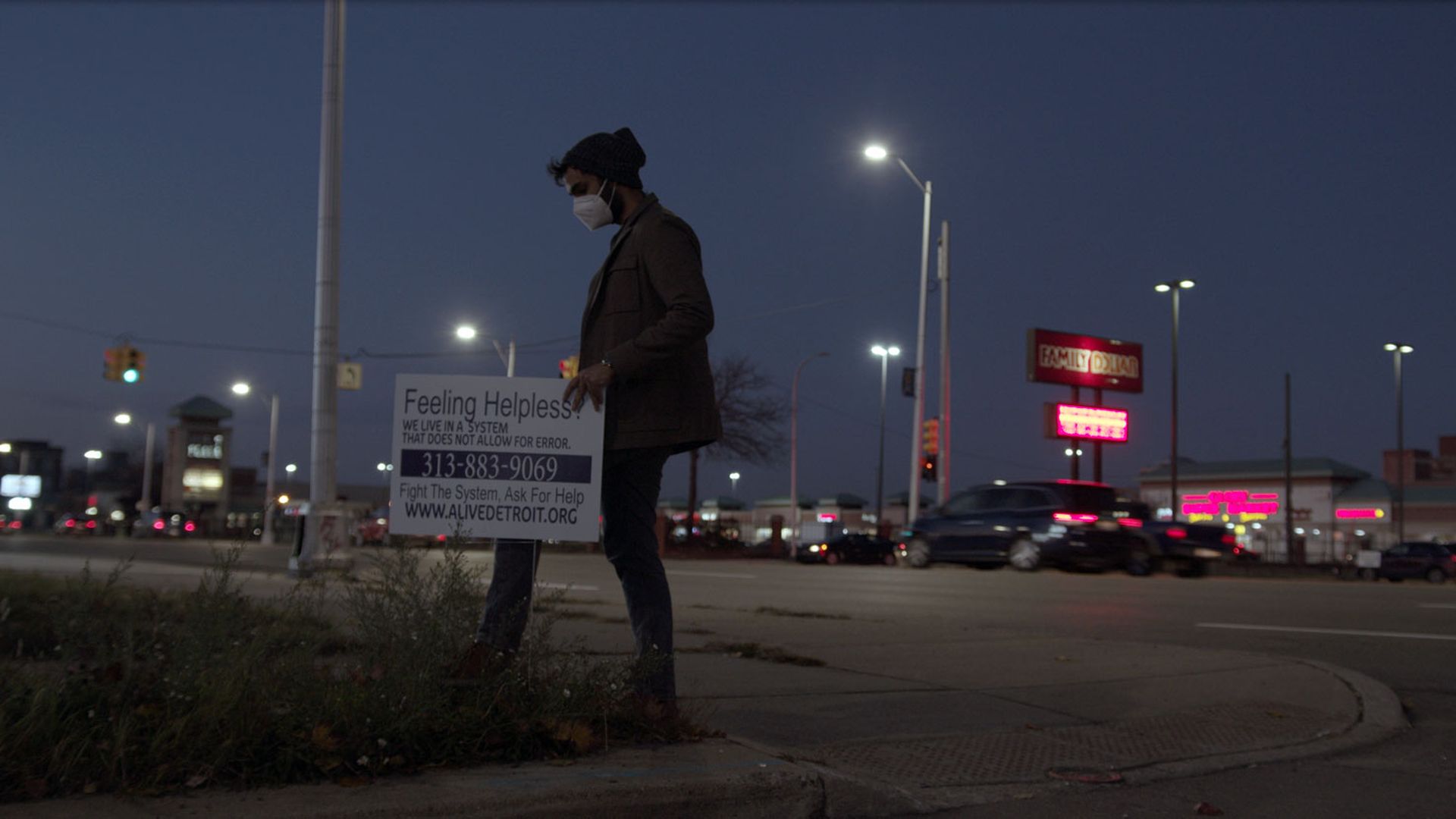 In nighttime, a person places a sign seeking people to reach out to Alive in Detroit. The person wears a beanie and a mask.