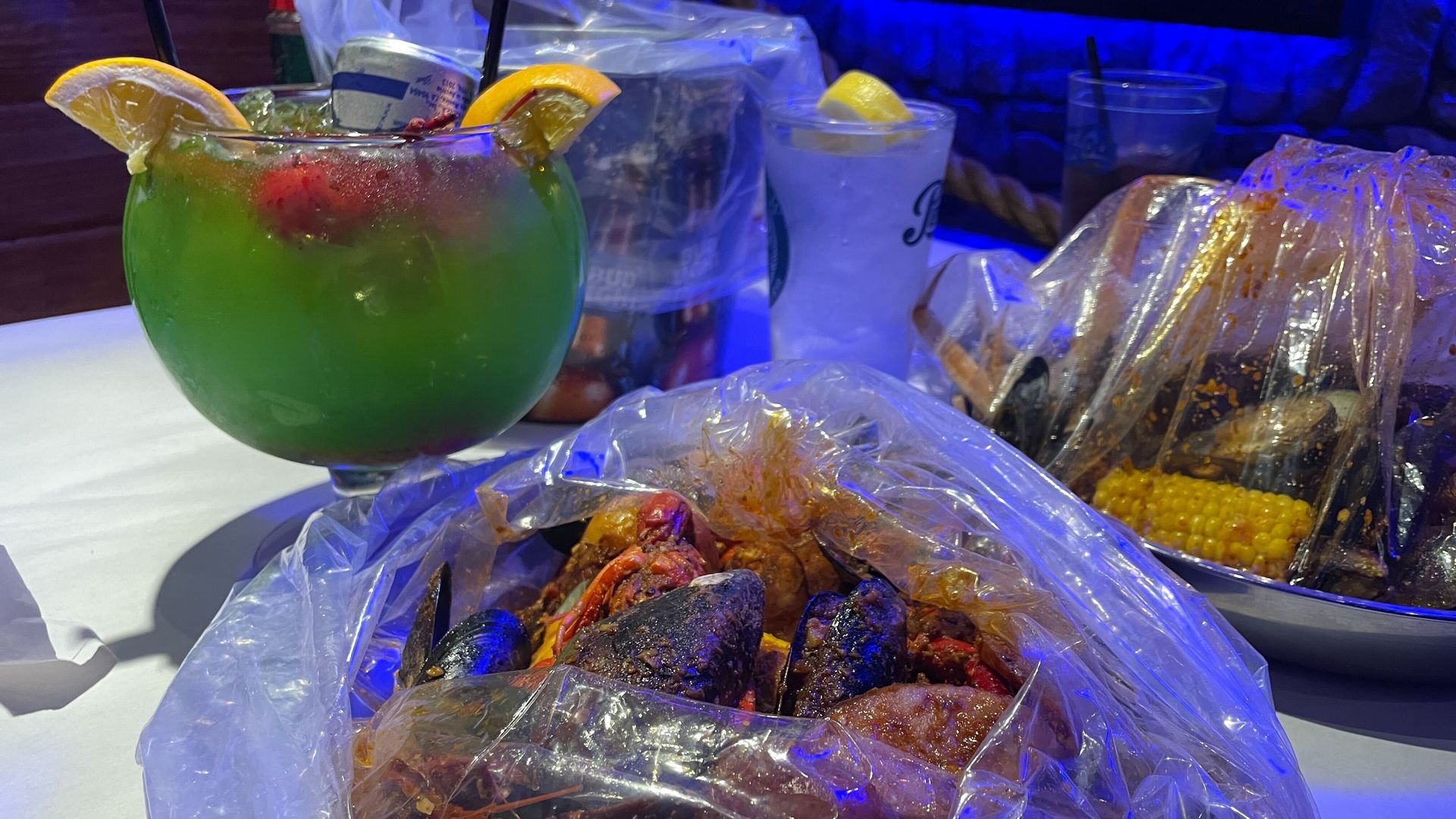 The lower center scene of this image displays a basket filled with seasoned crawfish, mussels, and potatoes. To the right of the basket is a large green fishbowl sized cocktail.