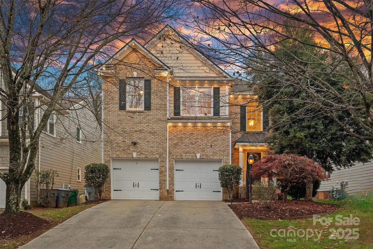 Two-story brick house with double white garage doors, black shutters, and lit porch at sunset; bare trees and landscaped front yard with a red bush and a lamp post.