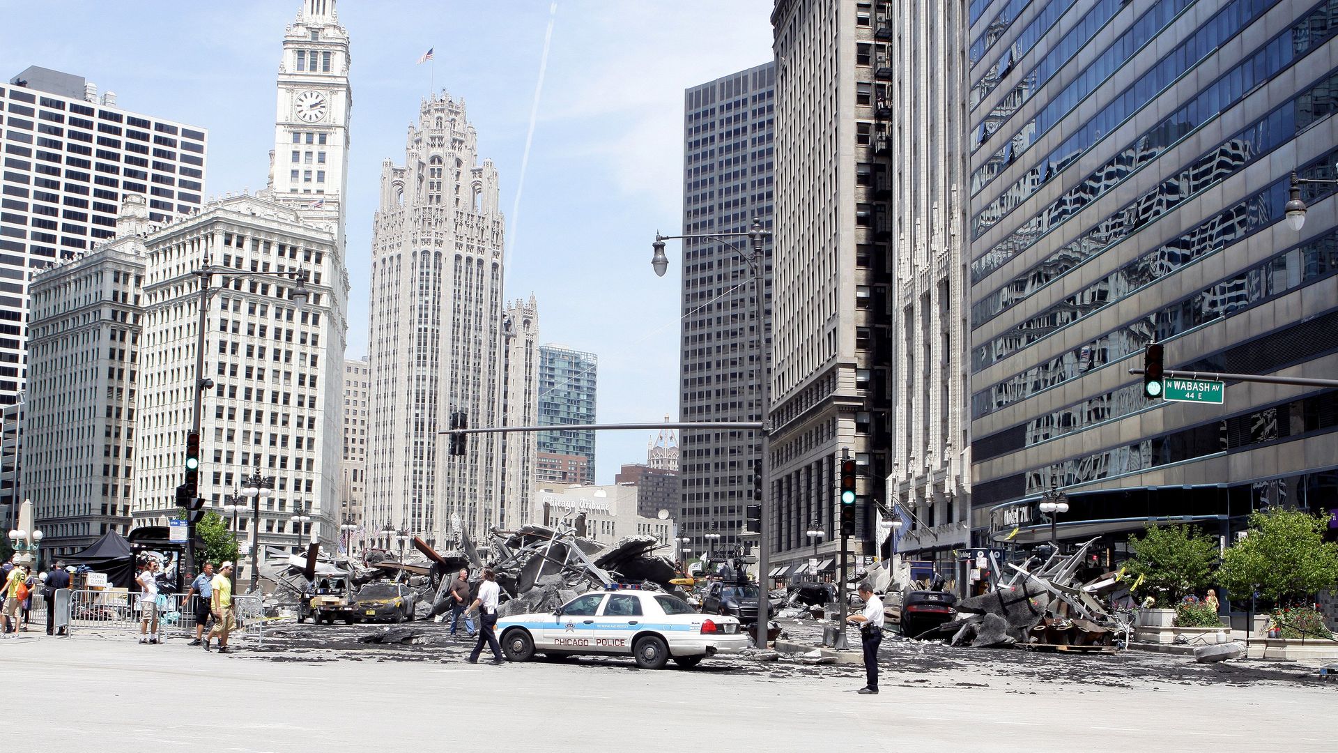 Chicago city street scene with a police car in the foreground, rubble across the road, pedestrians nearby, and tall historic and glass office buildings against a blue sky; N Wabash Ave sign visible.