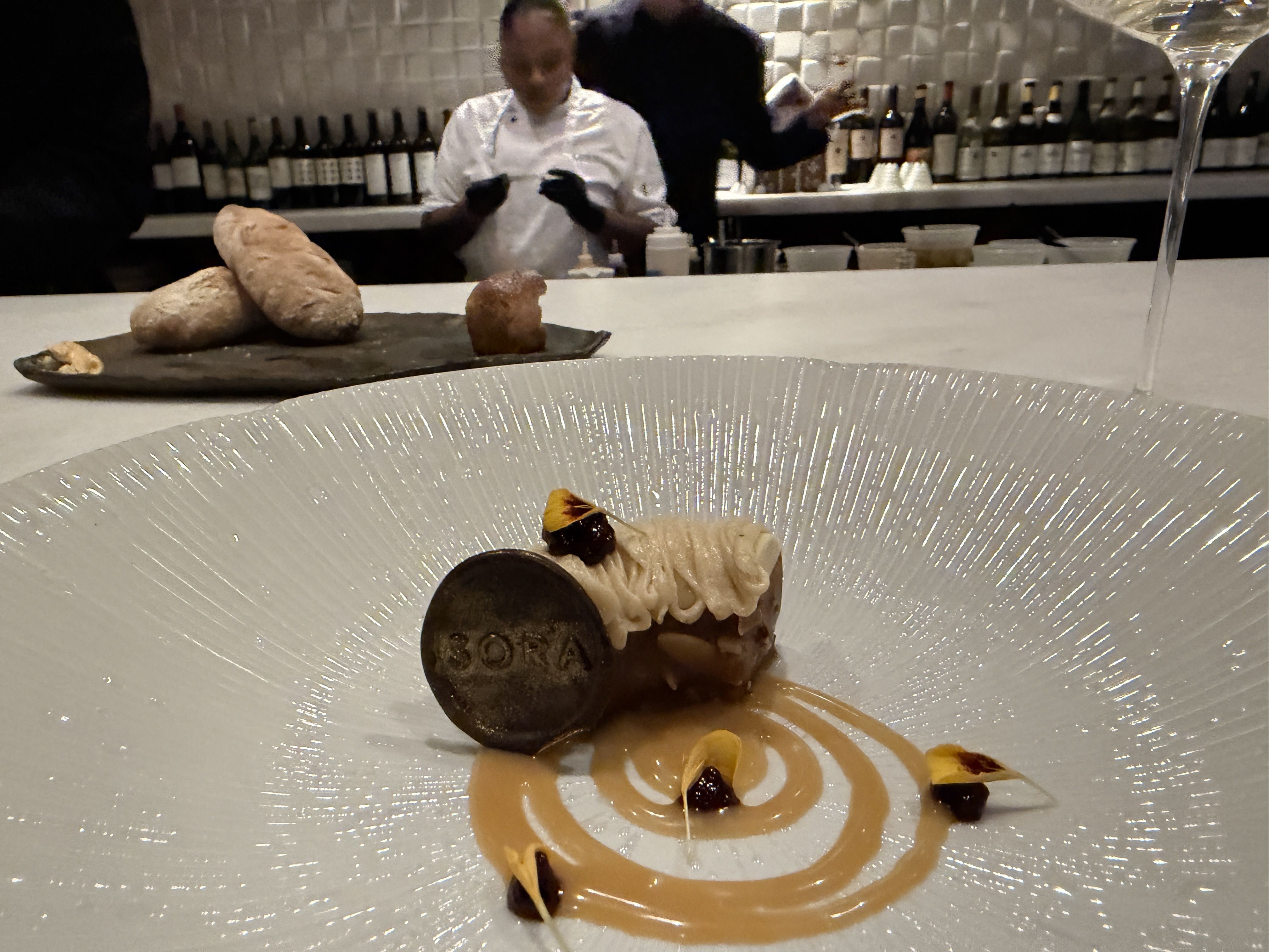 A plated dessert with a small cake topped with cream and a chocolate disk labeled "BORA" on a white decorative plate, with caramel sauce swirls, yellow petals, and dark dollops. Bread and chefs blurred in background.