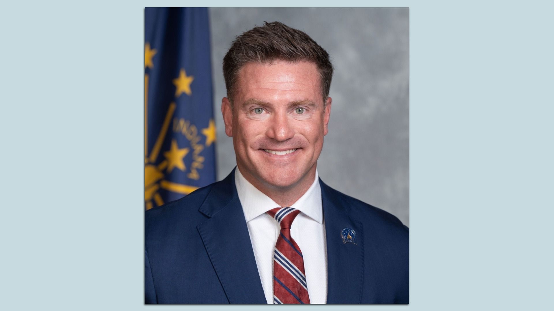 Portrait of a smiling man in a navy suit and red striped tie, with a blue flag and yellow stars behind him against a gray studio backdrop.