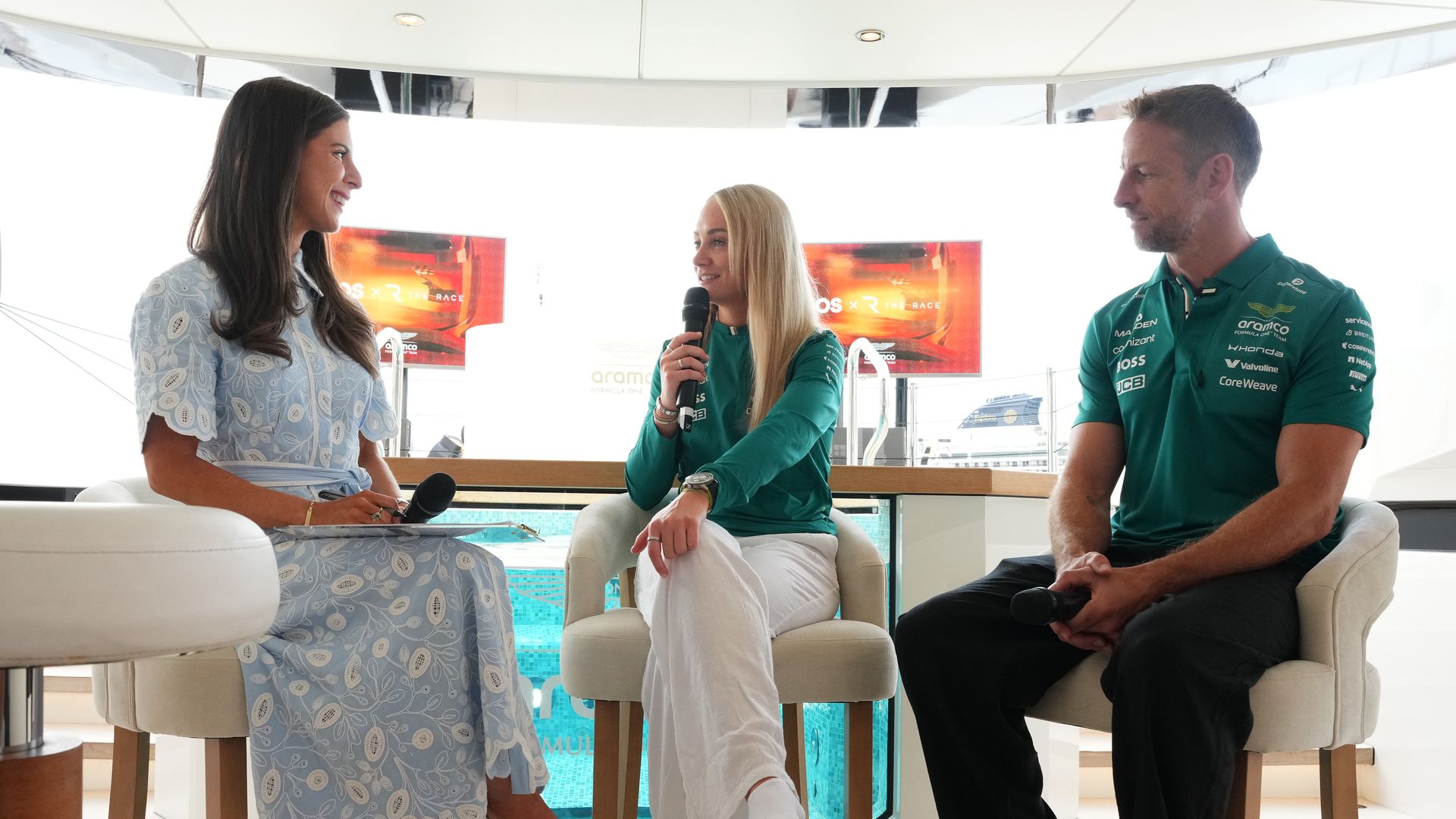 Three people sit on stools in a bright, modern yacht interior; left woman in a blue lace dress holds a microphone, middle blonde in teal speaks, right man in teal polo listens.