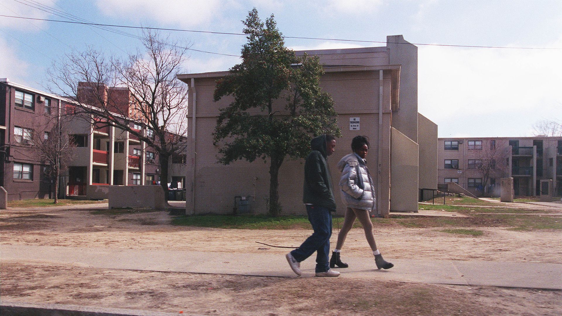 Two people walking on a sidewalk with an apartment complex in the back.