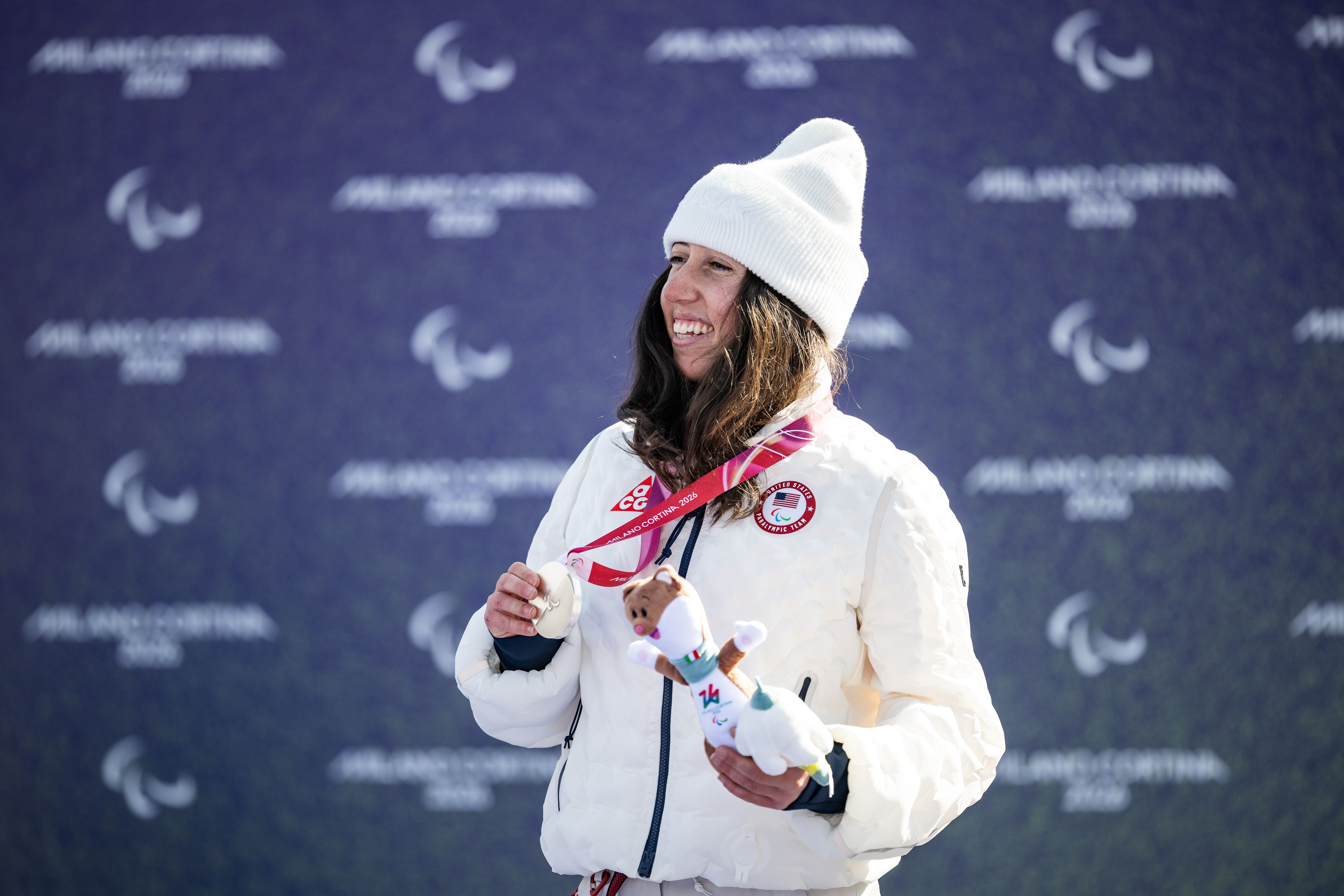 Kate Delson holds up her silver medal and a stuffed toy at the Paralympics while wearing a white Team USA jacket and white beanie.