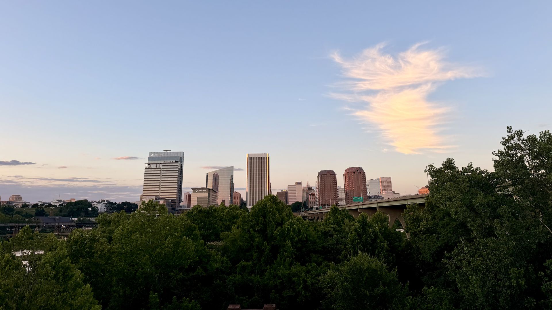 City skyline at dusk with tall buildings along the horizon, a highway overpass on the right, and dense green trees in the foreground beneath a blue sky with a pink-tinged cloud.