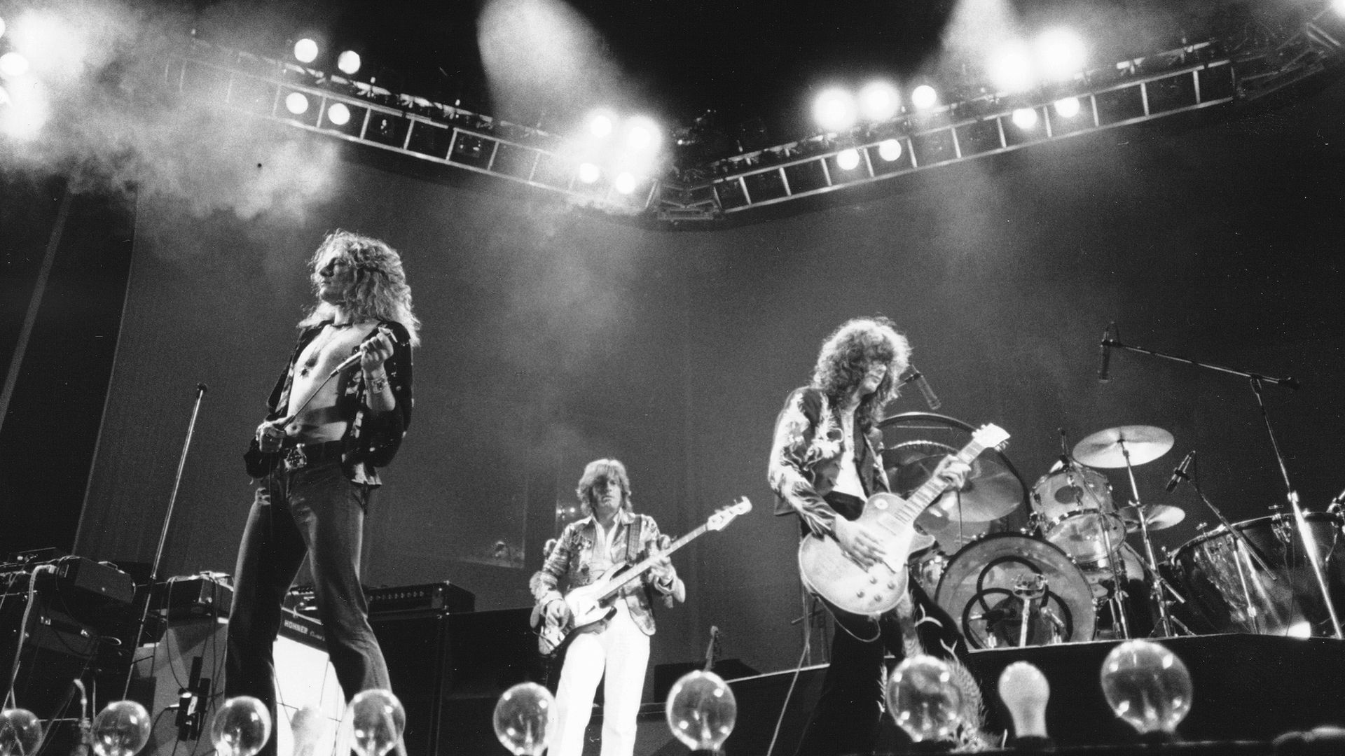 Black-and-white photo of a rock band performing on stage: shirtless, curly-haired guitarist left; bassist center; guitarist with a Les Paul right; drums in back, bright lights and smoke.