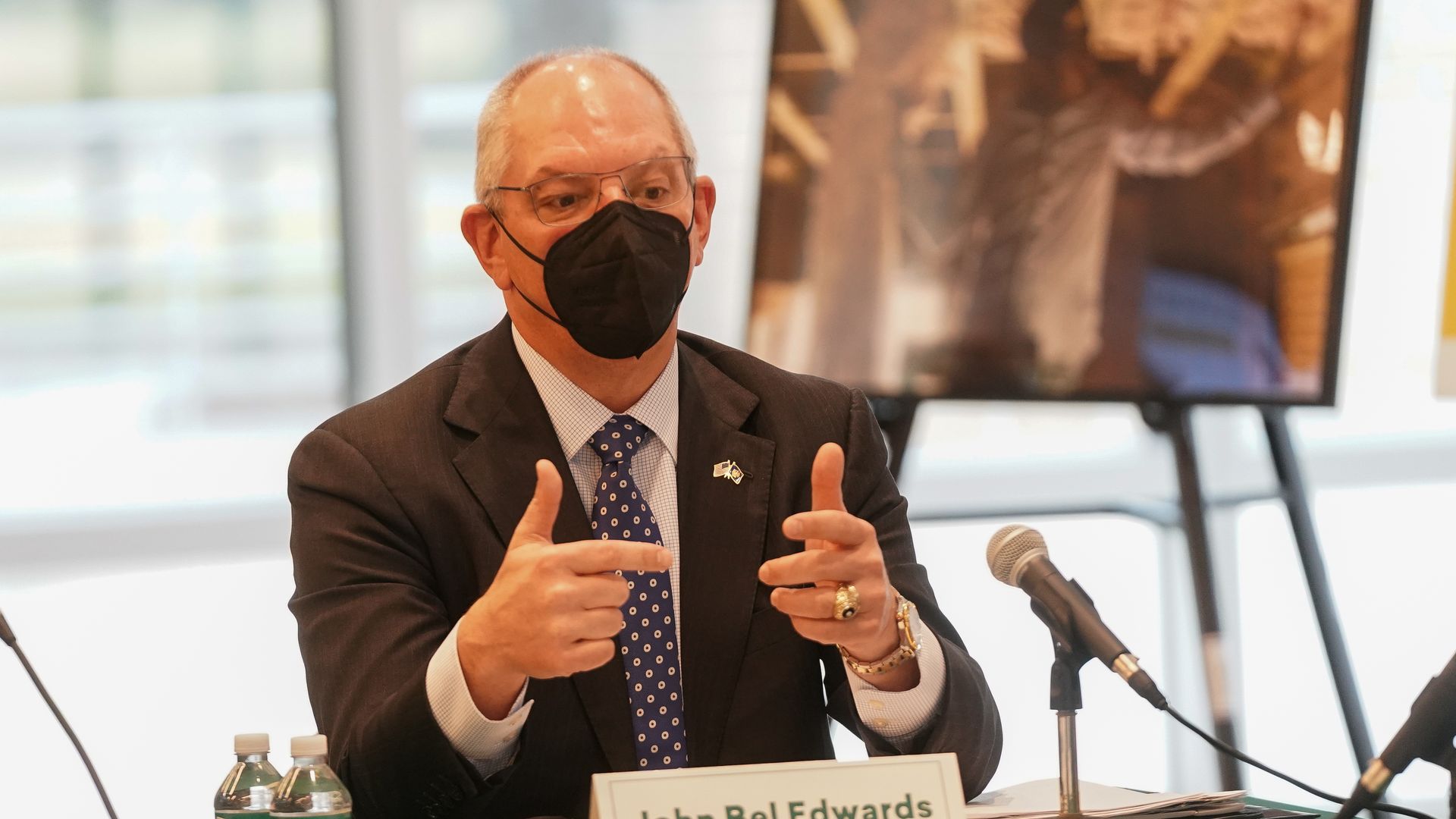 Photo of a masked John Bel Edwards sitting at a table and gesturing with his hands