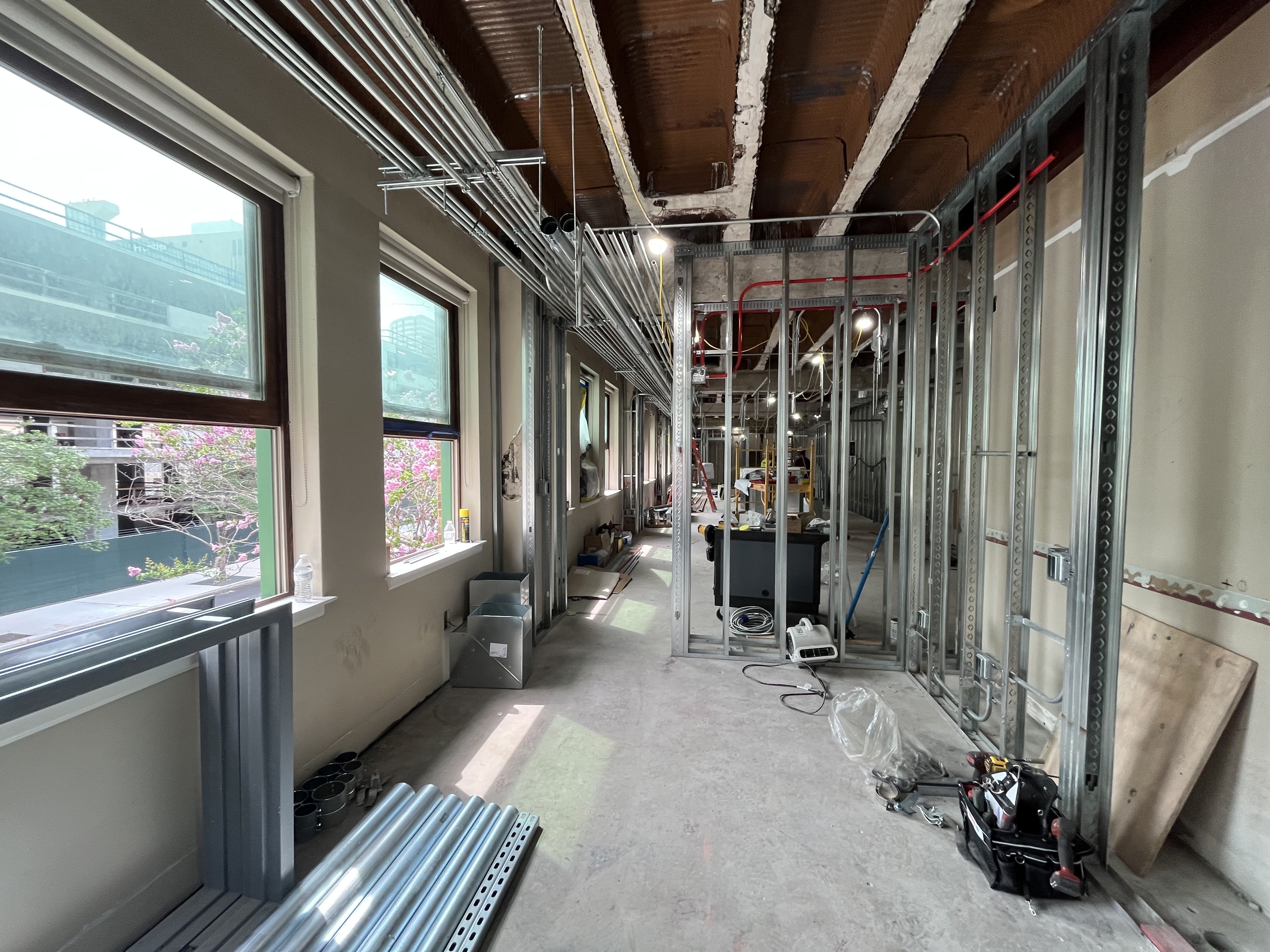 Indoor construction area with exposed metal stud framing, unfinished ceiling with wooden joists, exposed electrical conduits, and scattered tools; large windows show an urban exterior.