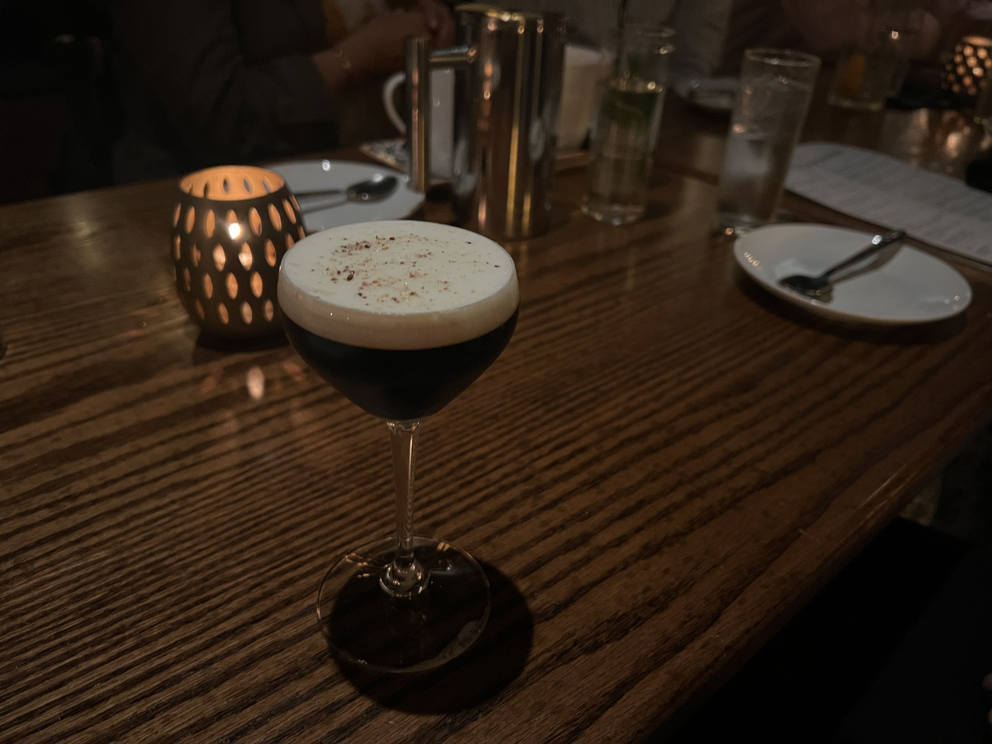 Dim restaurant table with a dark, foamy cocktail in a coupe glass in the foreground; a warm candle lantern and plates, cutlery, and tall glasses glow in soft light.
