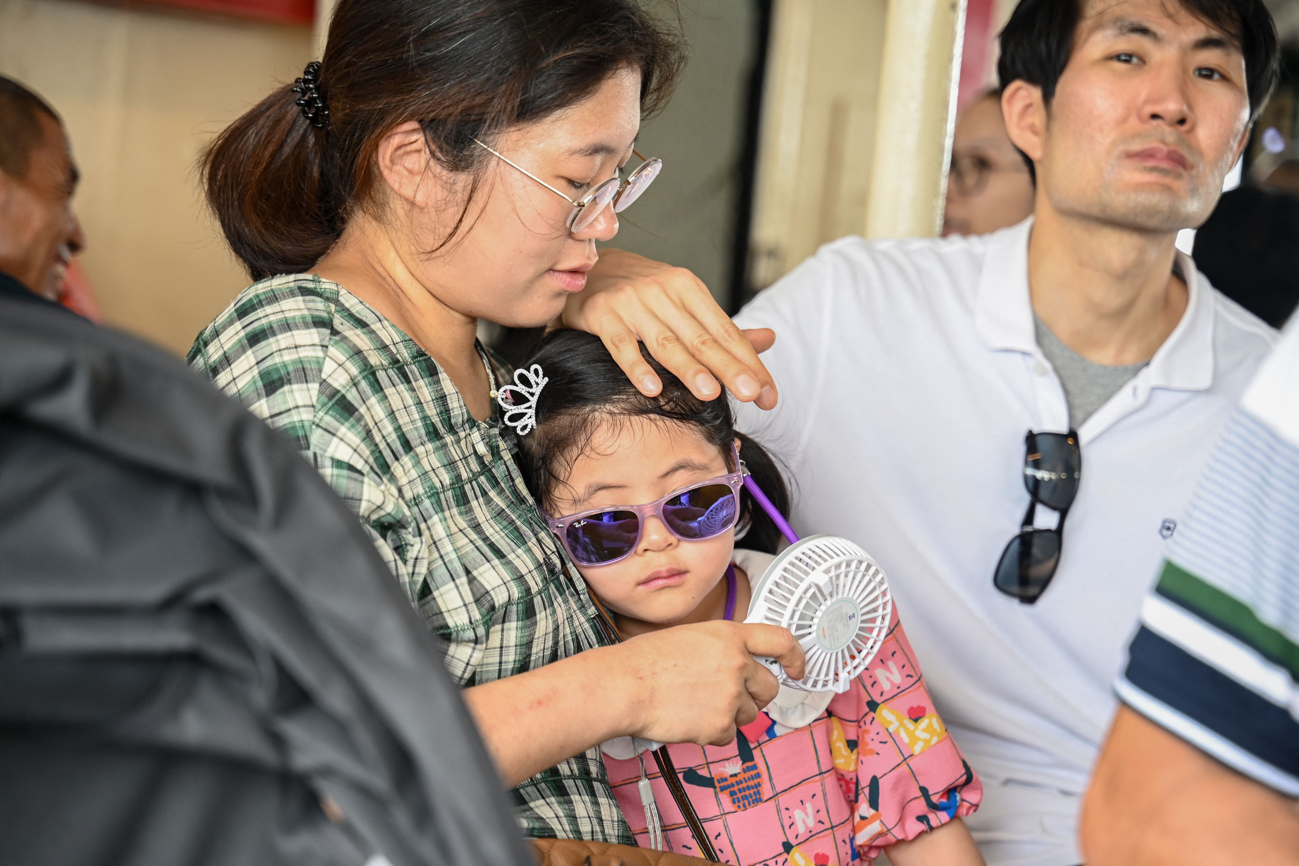 A girl being held by an adult who holds a small portable fan near her face. 