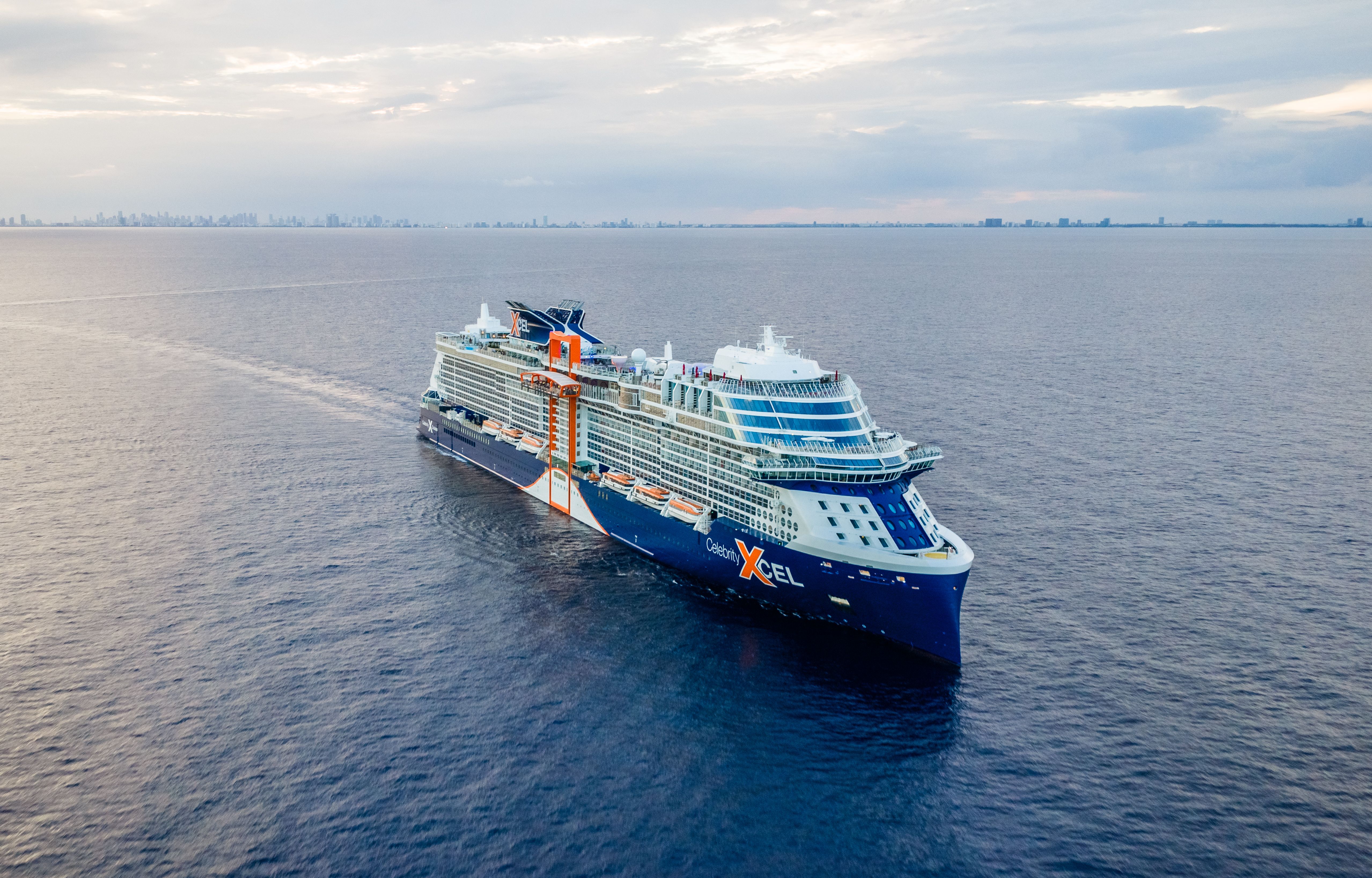 Large blue and white cruise ship with some orange in it. The ship is the new Celebrity Xcel ship show sailing on calm ocean waters with a distant city skyline under a cloudy sky.