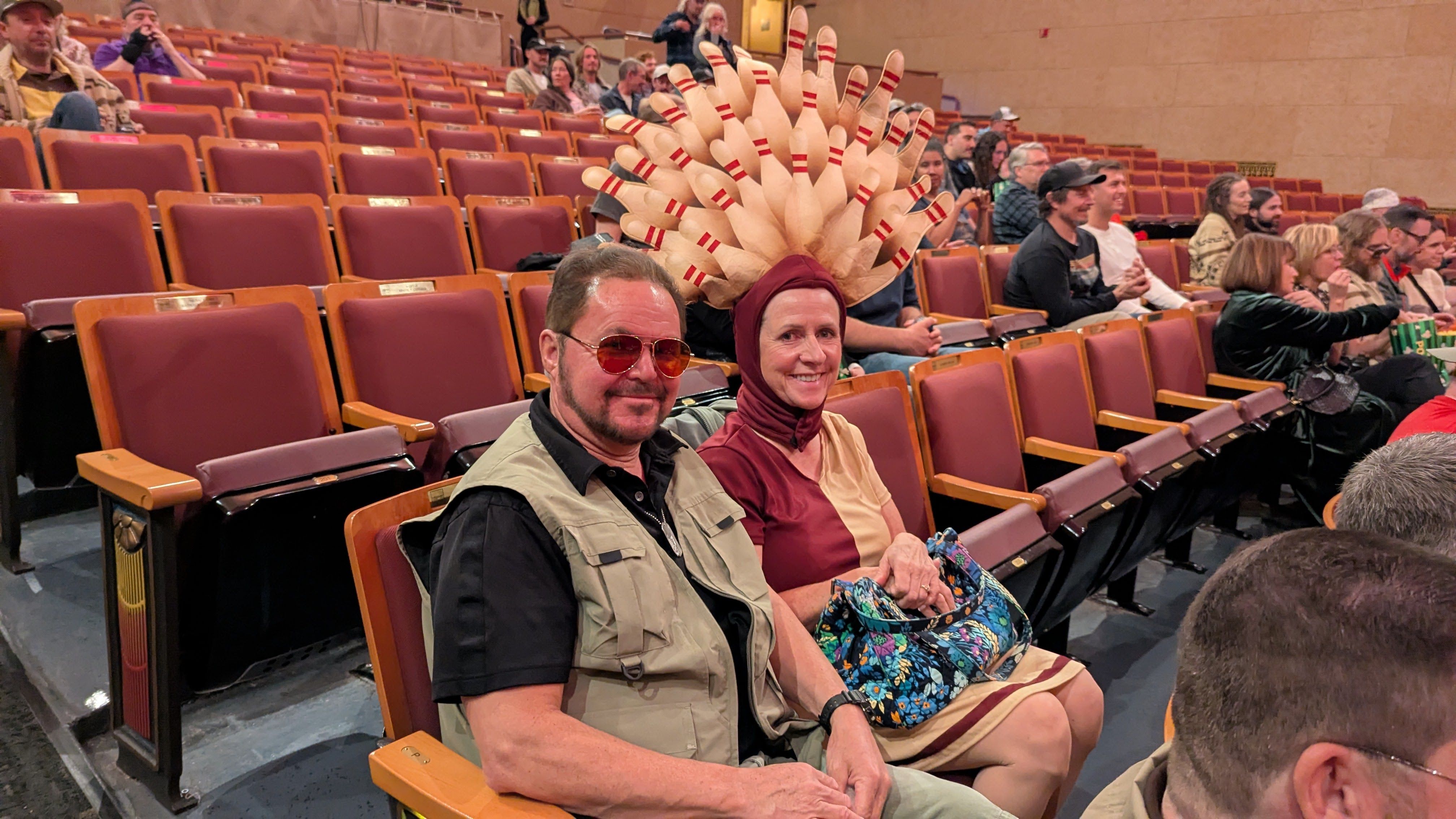 A man in a khaki vest and pants with a woman wearing a busby berkley style fan headpiece of bowling pins.