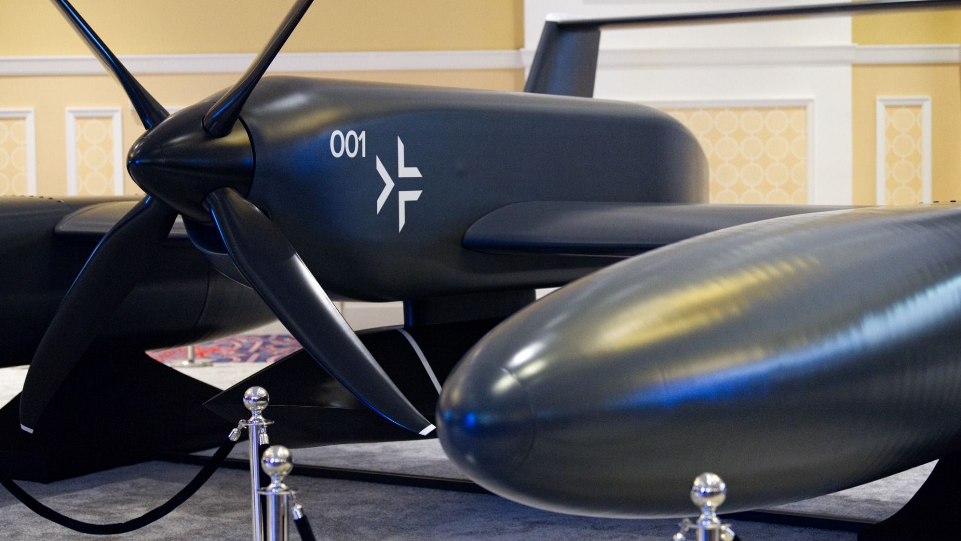 Close-up of a dark-colored aircraft nacelle with a propeller, marked 001 and a white geometric logo, displayed indoors against yellow patterned walls with white trim.