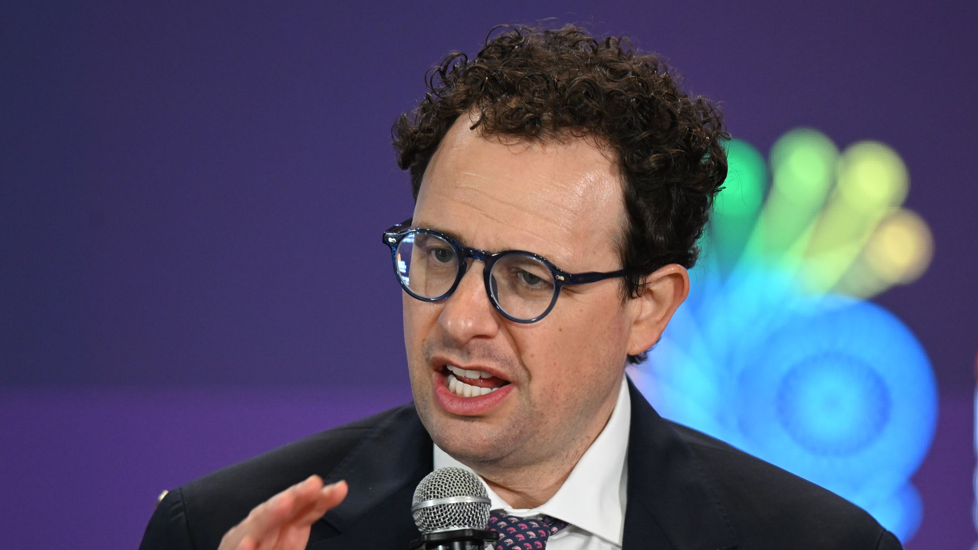 Anthropic CEO Dario Amodei, in a suit and tie and wearing glasses, holds a microphone and gestures with his right hand. 