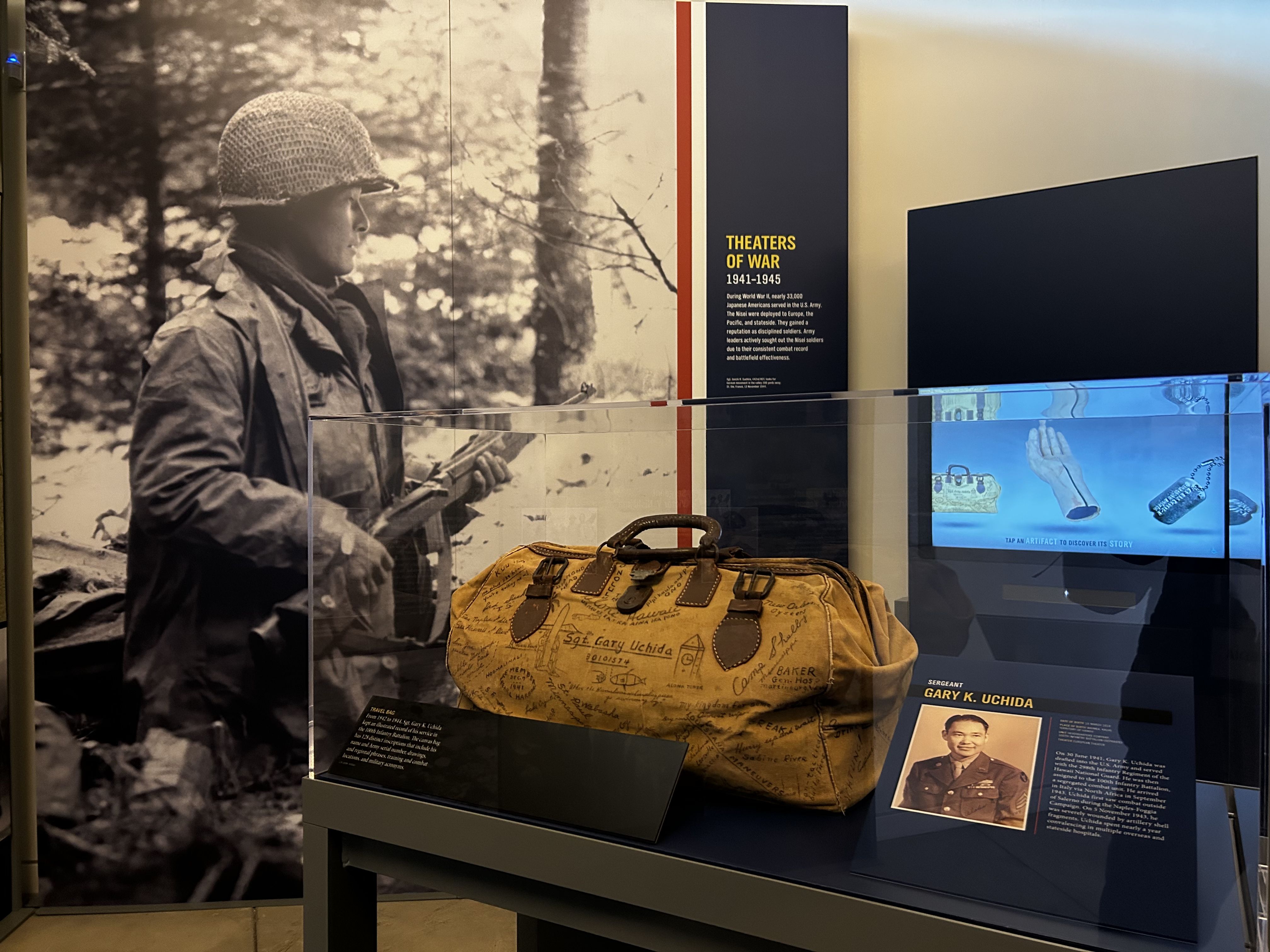 Museum exhibit featuring a yellow travel bag signed by Sgt. Gary K. Uchida, a soldier in WWII, with a black-and-white background photo of a soldier holding a rifle in a forest.