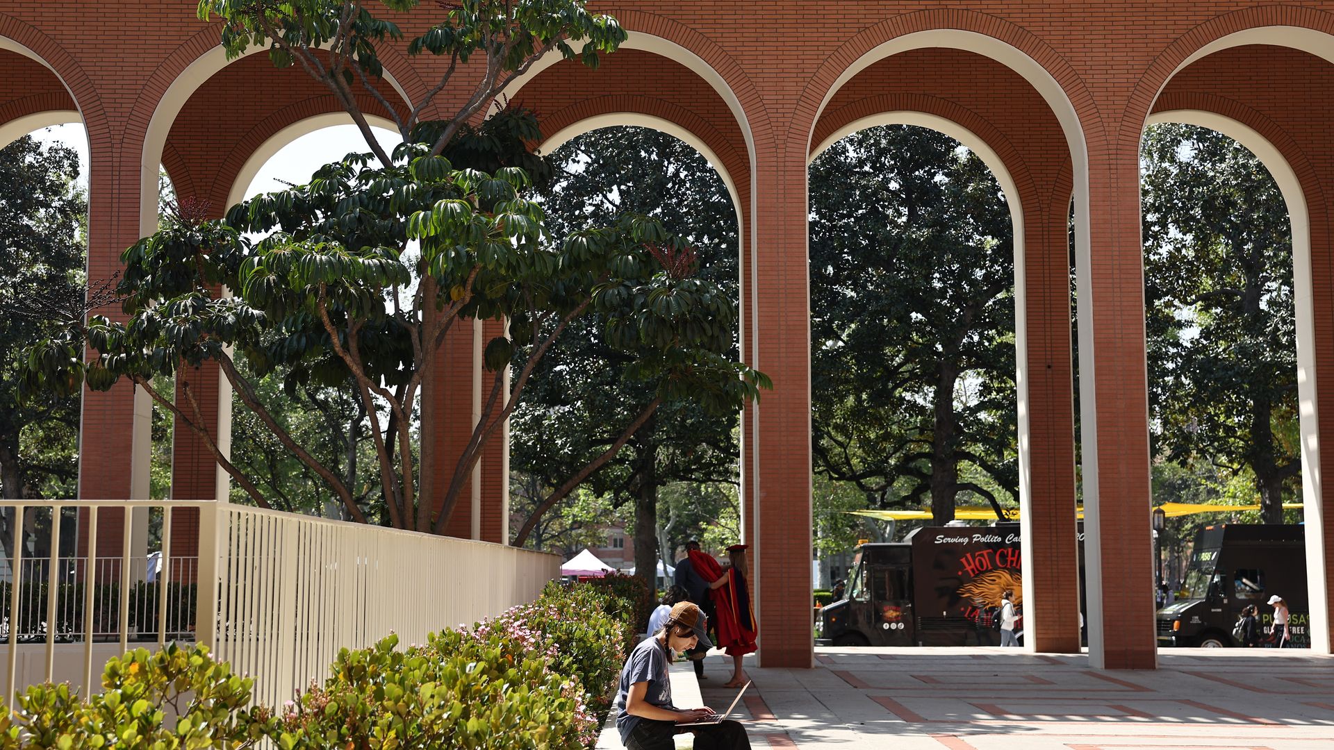 A student sits while typing on a laptop outdoors. Behind him, five arches are seen with many trees in the background. 