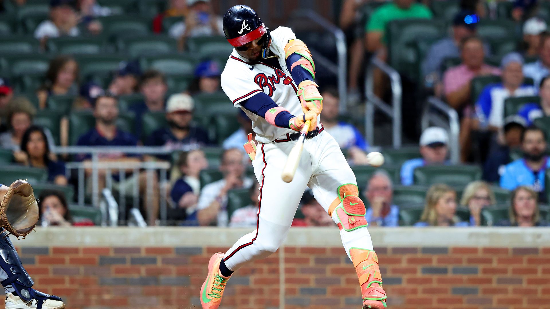 A player wearing a white Atlanta Braves uniform swings a baseball bat towards a baseball. Fans watch in the background and a catcher's glove and foot are also seen.