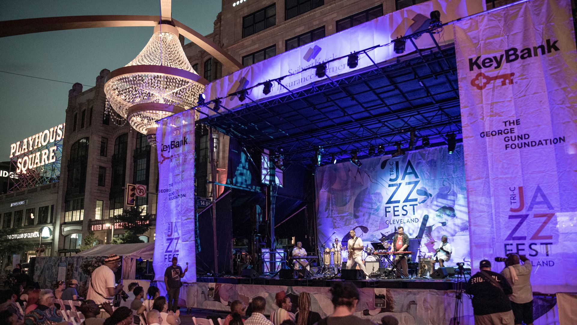 Nighttime outdoor concert at Playhouse Square. A lit crystal chandelier hangs above a stage draped with banners for KeyBank and The George Gund Foundation. Musicians perform as a crowd watches.