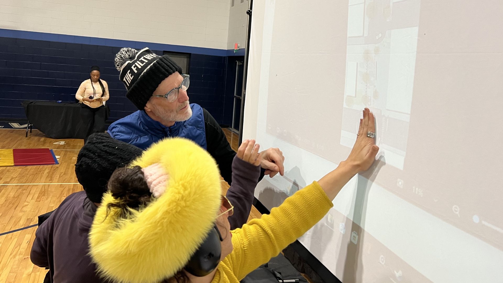 Several people stand indoors in a gym with wooden floors, interacting with a large white projector screen; one person in a bright yellow furry hat touches the screen as a man in a black beanie looks on.