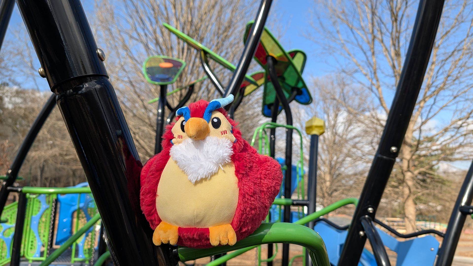 Red and yellow plush bird toy with white beard and blue eyebrows sits on green metal bar at colorful playground with bare trees under clear blue sky.
