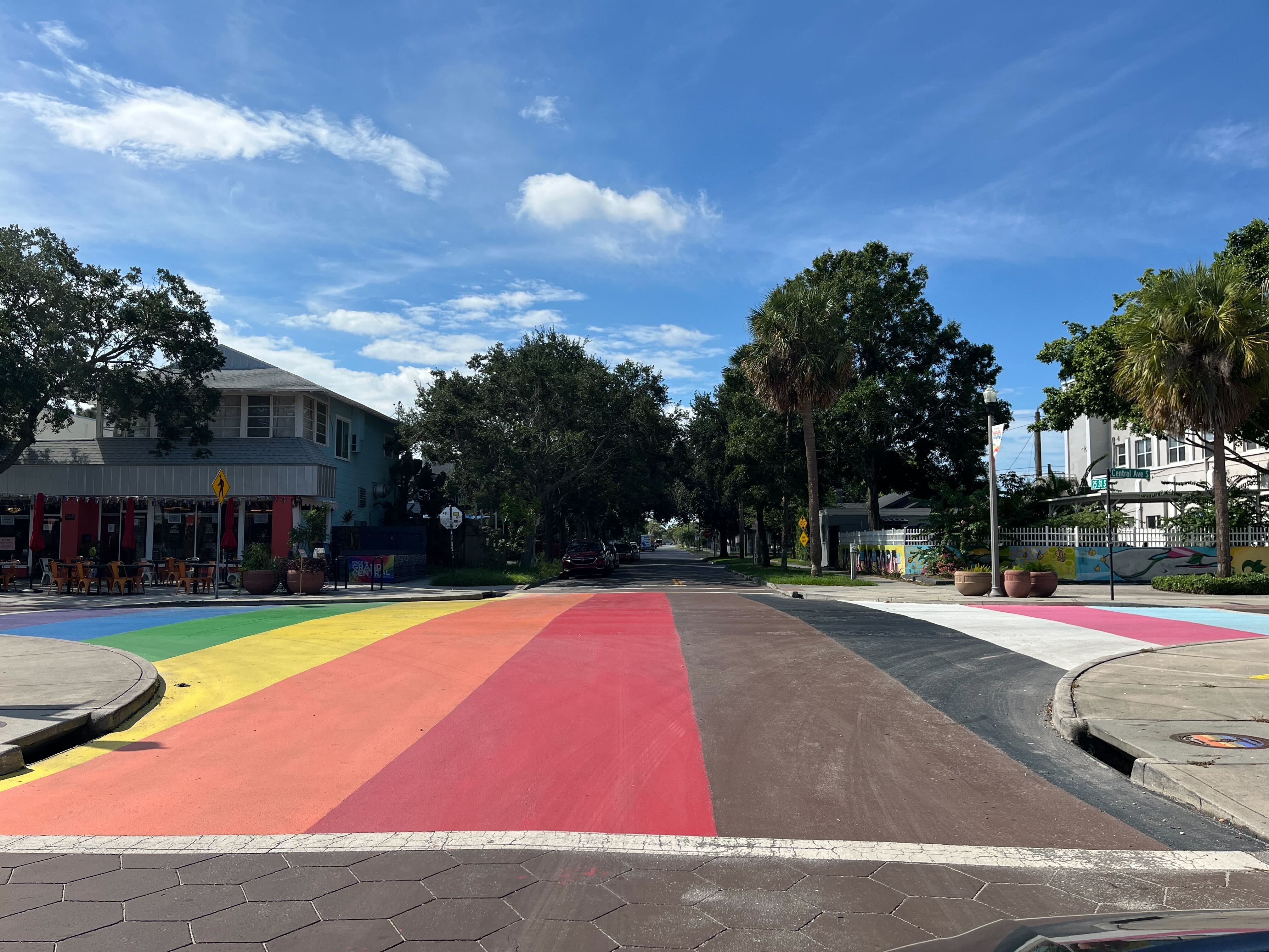 Street crossing painted in rainbow and transgender flag colors with trees, a building with outdoor seating, and a blue sky with clouds overhead.