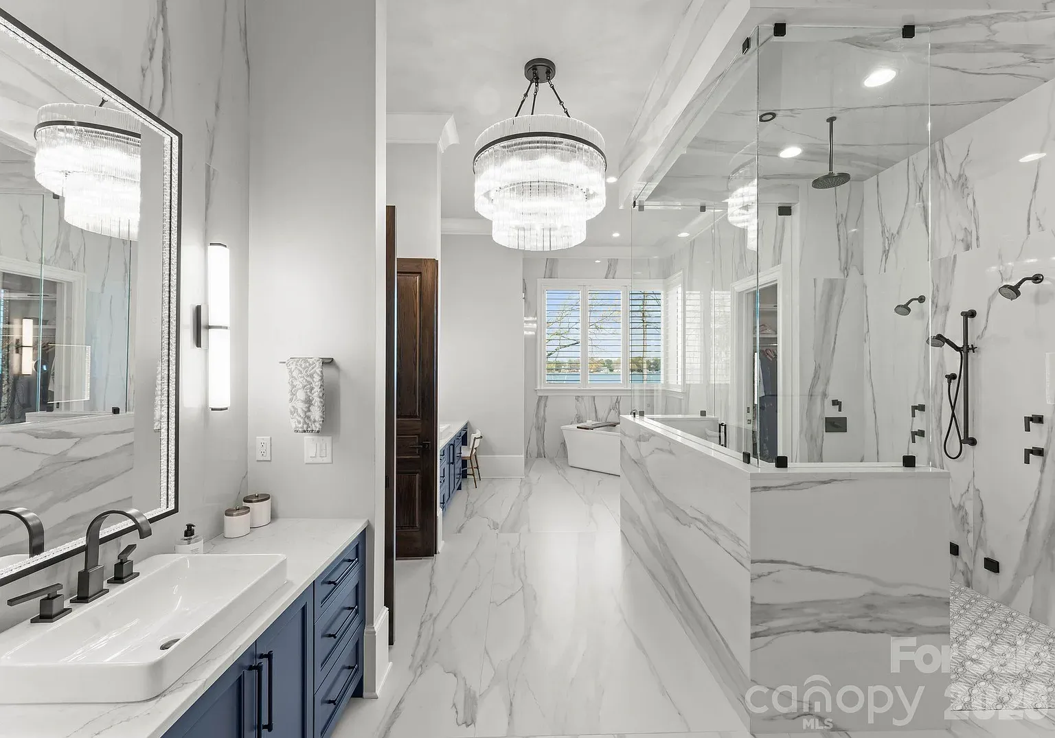 Luxurious marble bathroom featuring gray-and-white veining, a blue double-sink vanity, crystal chandelier, glass shower enclosure, and a freestanding tub by a window.