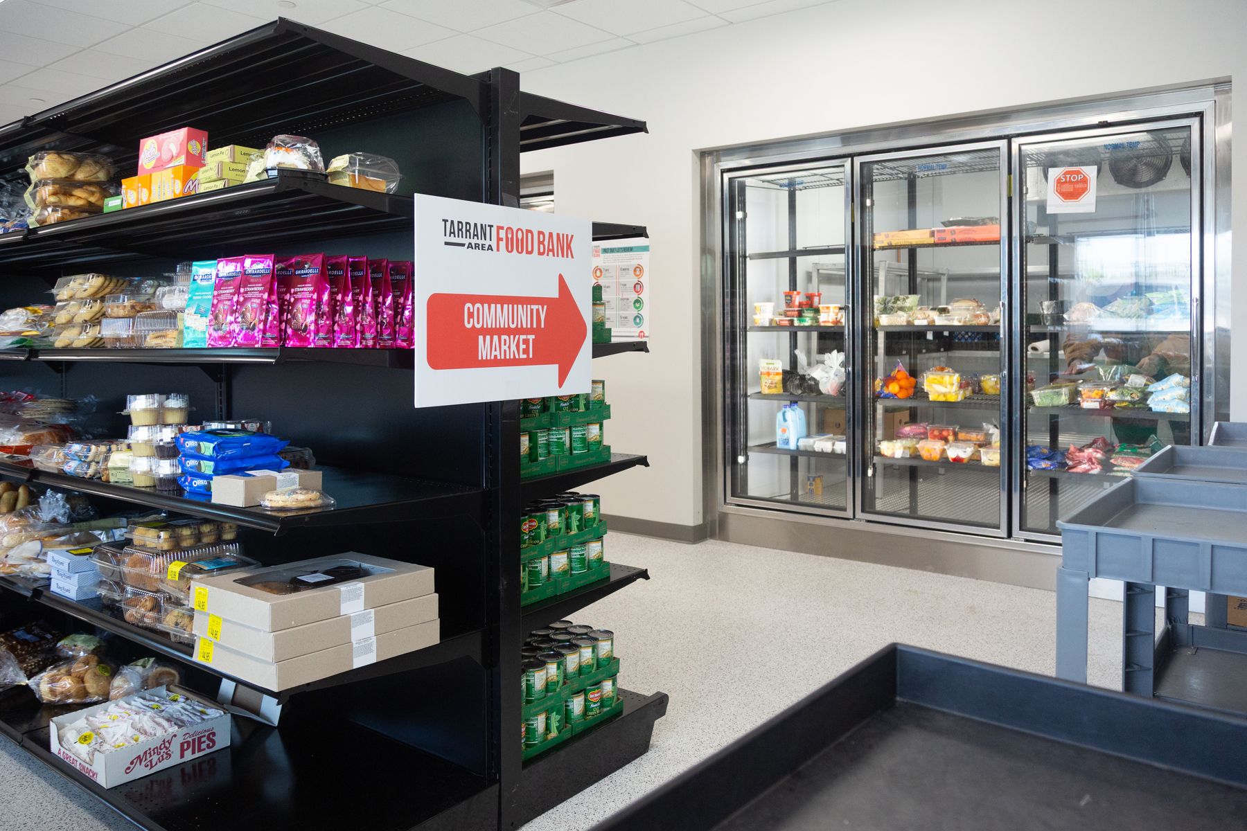 Shelves stocked with packaged bread, snacks, and canned goods next to a refrigerated section holding various fresh foods inside a bright community market at Tarrant Area Food Bank.