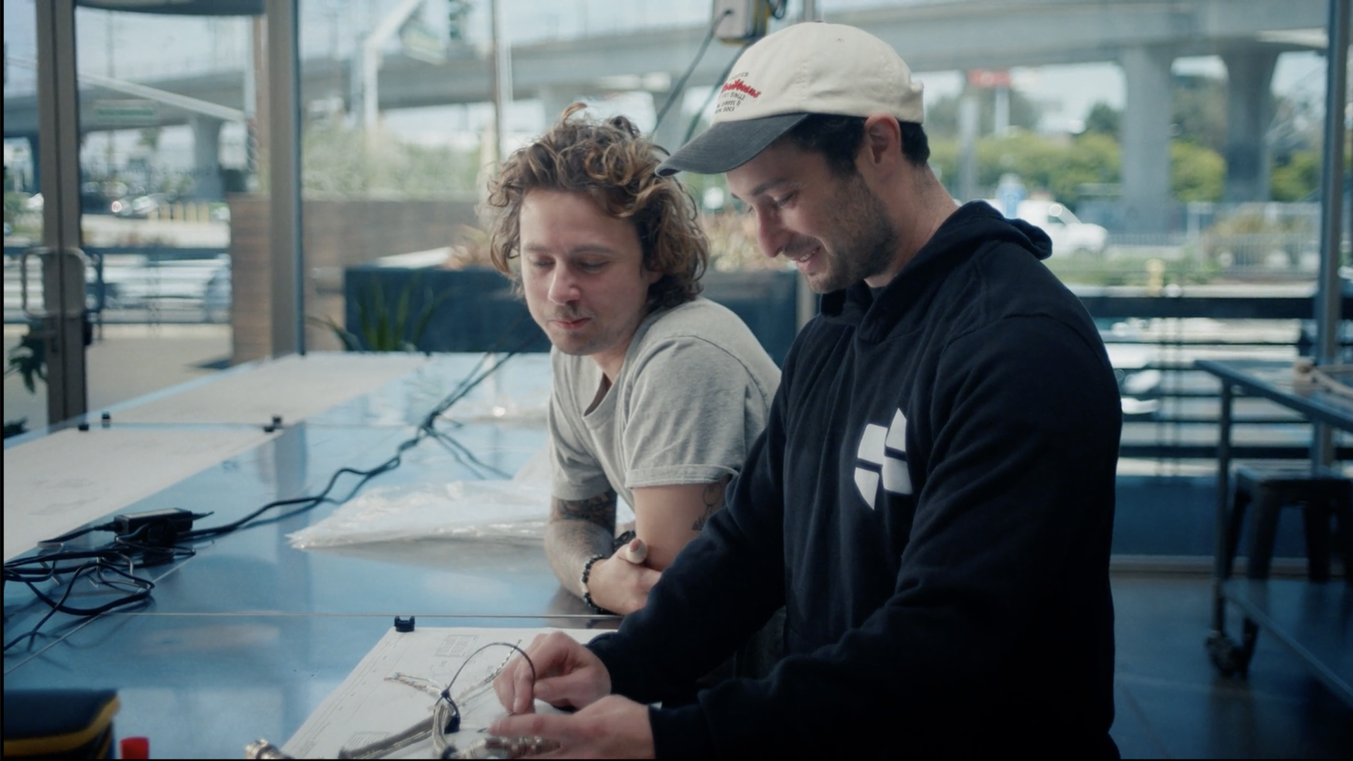 Two men, one in a hoodie and one in a tshirt, stand over a large desk. A bridge and a few cars can be seen through the windows behind them.